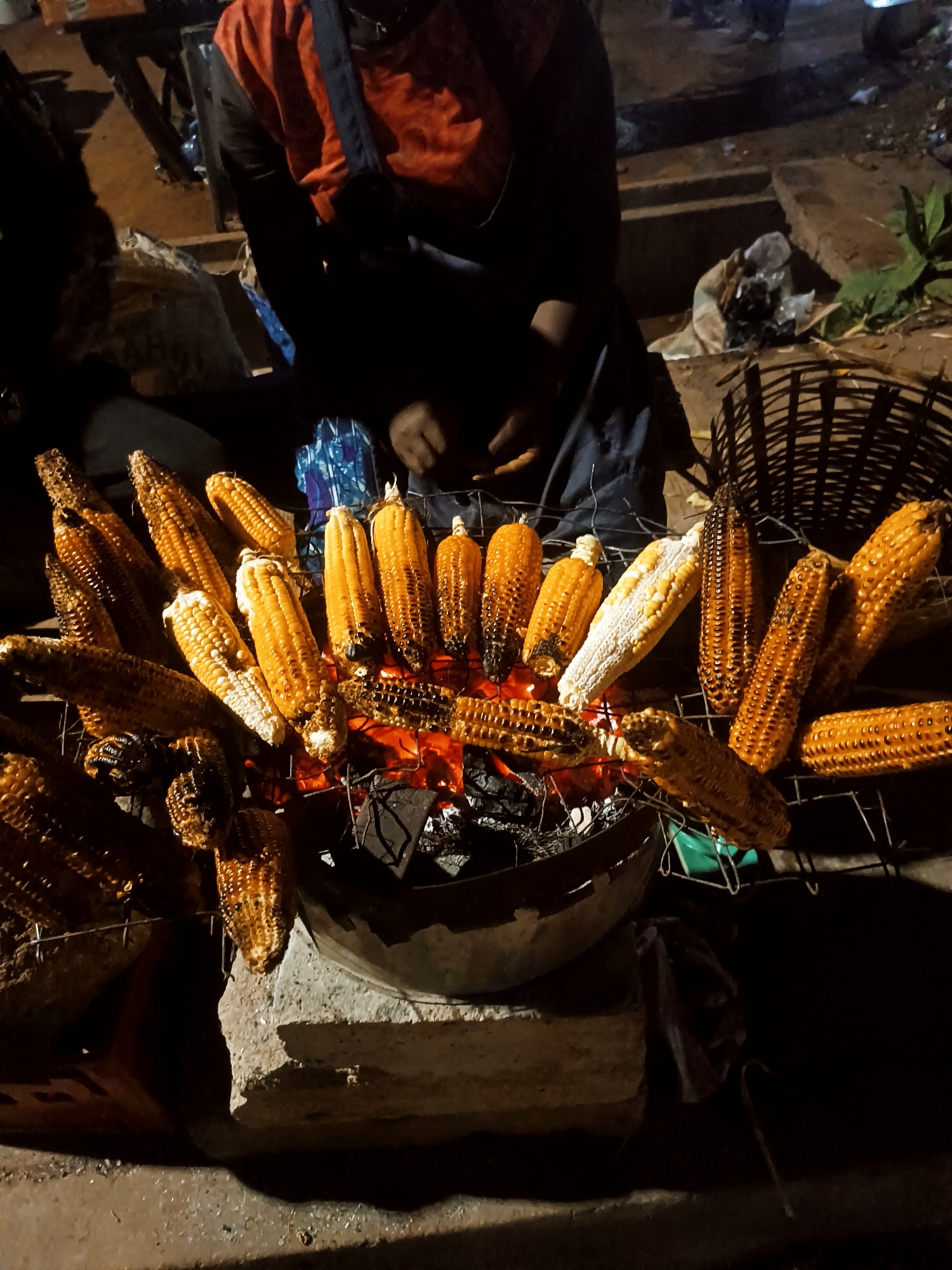 The image shows a street food vendor in Bukuru, Nigeria, grilling corn on a charcoal brazier at night. The vendor, wearing an orange top and dark apron, sits behind a grill laden with roasted corn, ranging in color from pale yellow to charred brown. The grill sits on a stone or concrete base. The foreground is dominated by the corn and the glowing embers of the charcoal fire. A woven basket is to the right of the image. The background is dark and shows indistinct figures and objects, indicating a busy street scene.