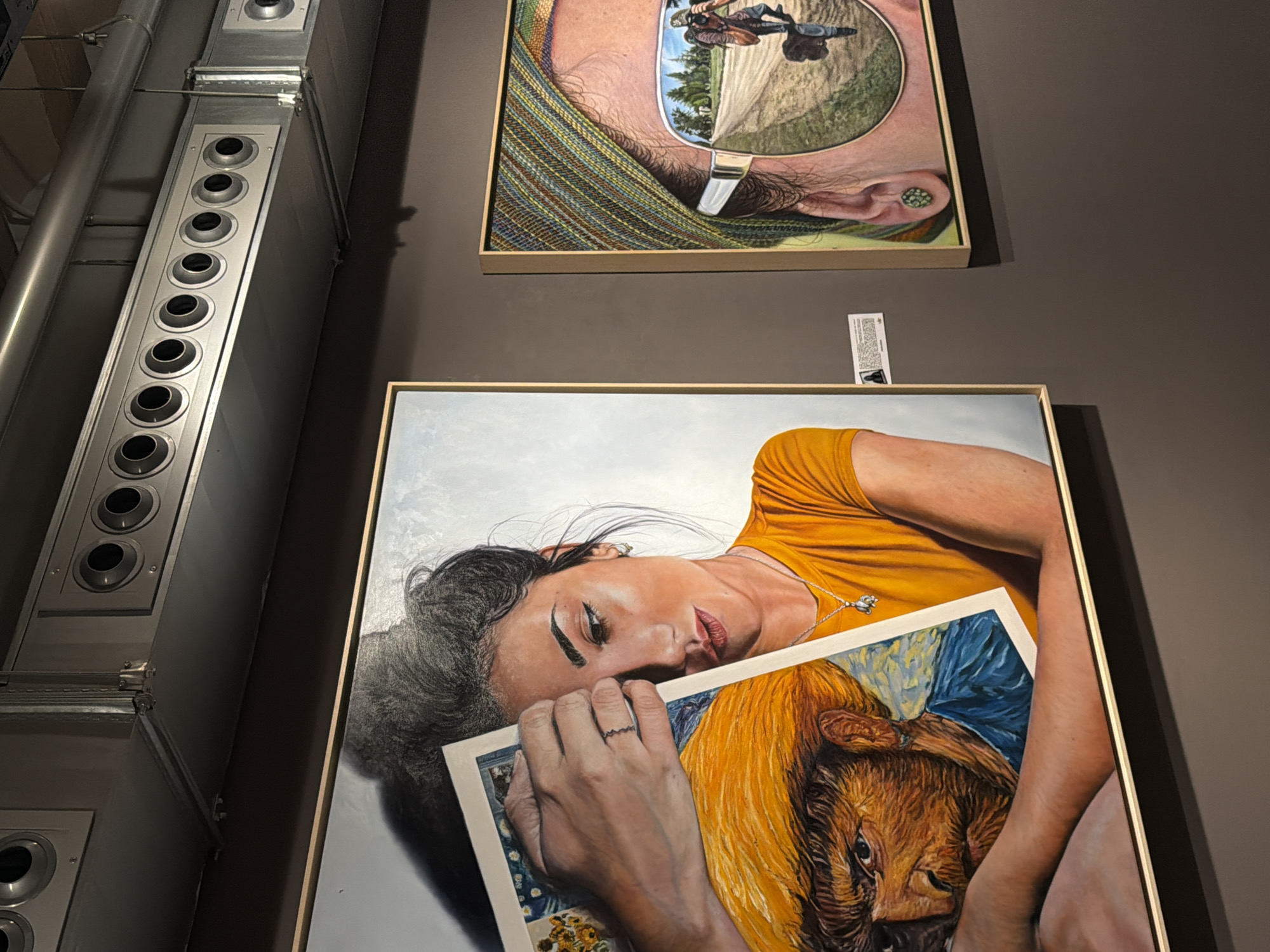 The media file displays two framed artworks hung on a textured brown wall. To the left, a large silver ventilation unit runs vertically, featuring a row of circular vents. The bottom portion of the ventilation unit is visible.
The upper artwork is a painting depicting a person's head and shoulders, seen from the nose upwards. The subject wears large, reflective sunglasses which mirror a landscape with a dirt path, trees, and what appears to be a person on a motorcycle. The person in the painting is also wearing a colorful, striped scarf around their neck. The painting is framed in a light-colored wood.
The lower artwork is a larger, portrait-oriented painting of a woman lying down, wearing a bright orange shirt. Her head is tilted back, and she holds a smaller painting or print in front of her chest. This inner artwork shows a close-up of a calf or young cow with a blue and yellow background. The woman has dark hair, defined eyebrows, and is wearing a ring with a zig-zag pattern on her finger and a delicate necklace with a charm. The painting is framed in a similar light-colored wood. A small white tag with some printed text is attached to the wall between the two artworks, slightly to the right of center. The overall setting appears to be an art gallery or exhibition space. No specific time of day or weather conditions are discernible. The emotions conveyed by the subjects in the paintings are ambiguous.