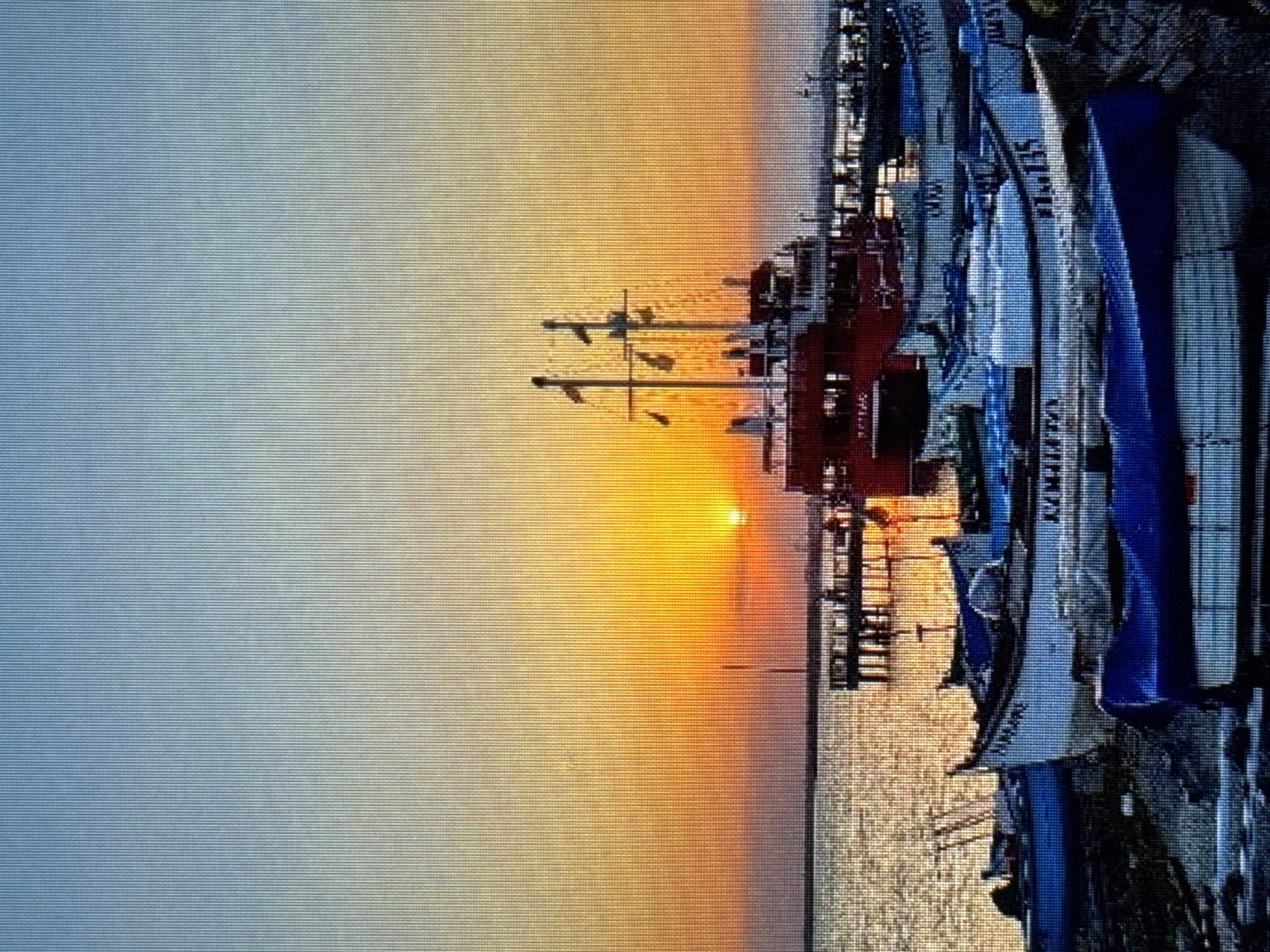 An evening scene depicts a harbor with a large red boat and several smaller fishing boats. The sun is setting on the horizon, casting a warm glow across the sky and water. A pier extends into the water, with a few figures visible on it. The smaller boats are lined up on the shore, some covered with blue tarps. The water is calm, reflecting the colors of the sunset. The overall impression is one of peace and tranquility.