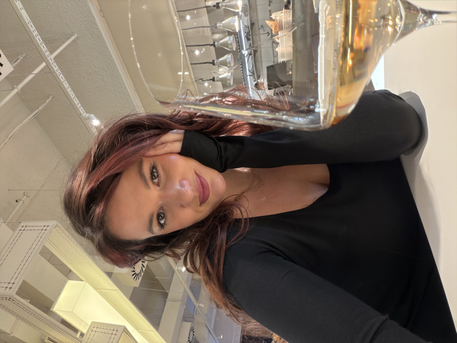 The image displays an adult female seated at a light-toned table. She has reddish-brown, wavy hair and is dressed in a black long-sleeved top with a low neckline, accessorized with a gold-toned layered necklace. Her right hand rests against her jawline, and she is looking towards the camera with a slight smile. In the right foreground, a wine glass filled with an amber liquid is partially visible. The background reveals a modern restaurant environment featuring an open kitchen. Within the kitchen, stainless steel surfaces, multiple metallic pendant task lights, and a partially visible individual in a white uniform are present. The ceiling above the dining area is fitted with distinctive rectangular, fabric-wrapped light fixtures exhibiting visible stitching patterns, alongside exposed ventilation ducts. This establishment is identified as Magaza Gastronomy Bar, located at Karađorđeva 2-4, in the Beton Hala area of Belgrade, Serbia.