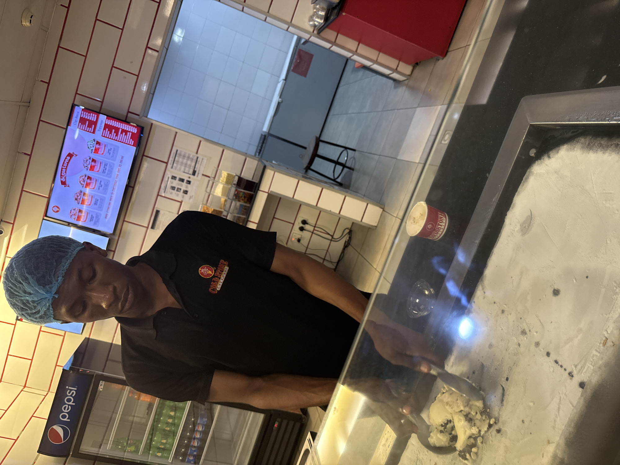 The scene depicts a man in a blue hairnet and a black polo shirt with a "Cold Stone Creamery" logo, preparing ice cream on a cold stone surface behind a glass display. A silver spatula is in his hand, mixing the ice cream. There is a cup labeled "Cold Stone" near the edge of the counter. Behind him, a Pepsi-branded refrigerator filled with beverages is visible. A digital menu board displaying ice cream flavors is on the wall. The location appears to be a Cold Stone Creamery in Abuja, Nigeria.