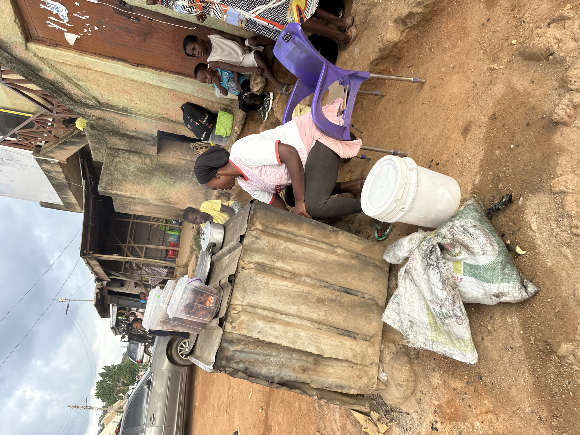 In Gwong, Nigeria, a woman wearing a pink and white shirt and a dark headscarf kneels beside a makeshift stall, preparing food. Her focus is on a large white bucket in front of her. Behind her, several people are visible. A young boy in a blue shirt sits on the ground, while other individuals are partially obscured. The scene is outdoors, with a rough, sandy ground and a simple, weathered structure that serves as a stall.
Several objects are scattered around the area: a purple plastic chair, bags of produce, and clear plastic containers filled with food. One of the bags, partially opened, displays green and white lettering. The overall impression is of a bustling marketplace or street-side vendor operation. The lighting suggests it's daytime, possibly late morning or afternoon. The sky is overcast, indicating cloudy weather. The activity is centered around food preparation and likely sales. The presence of people of various ages suggests a community gathering or a typical day in a busy commercial area.