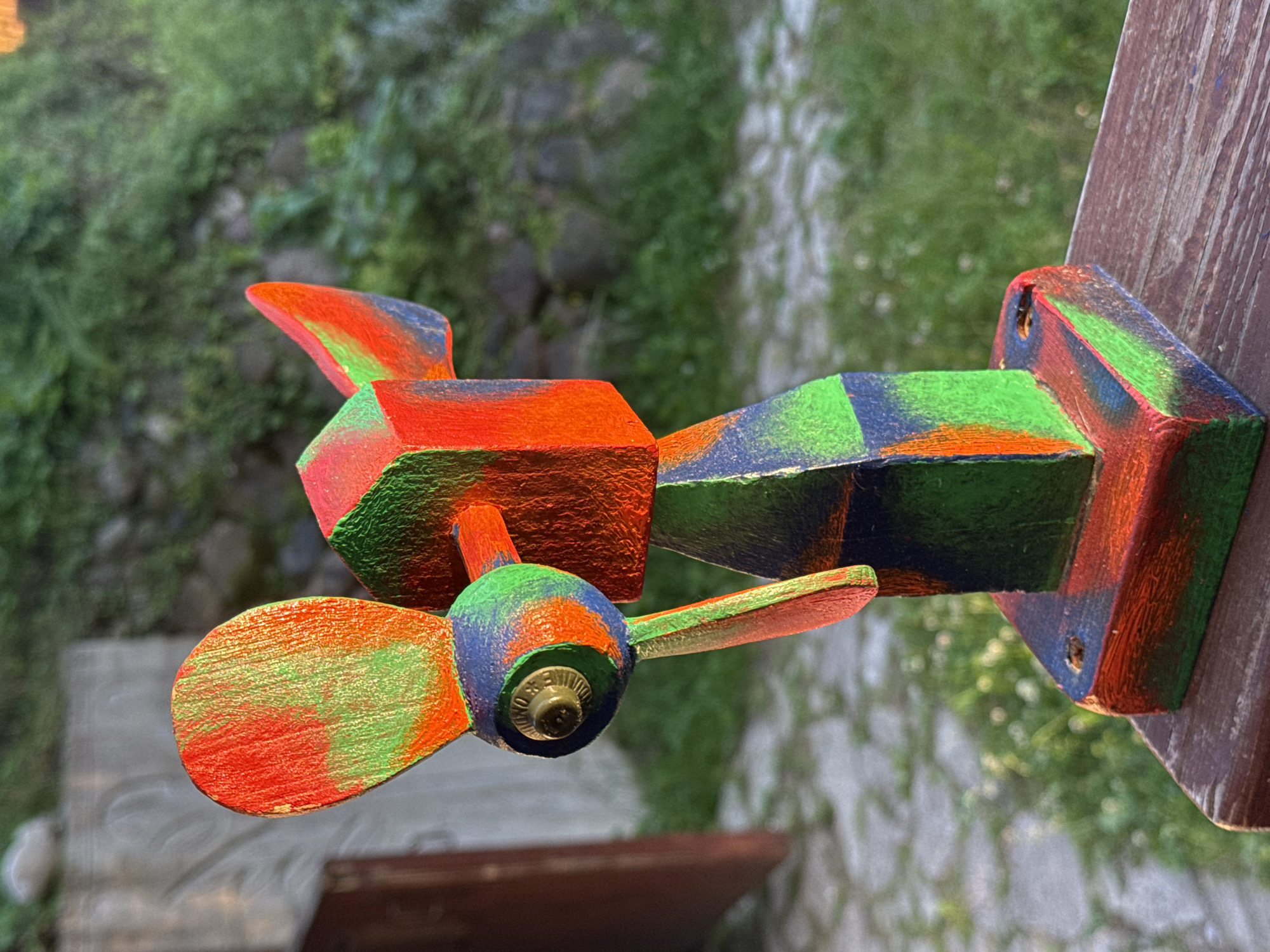 The image shows a brightly painted wooden airplane toy, with a red, green, and blue color scheme, attached to a wooden post. The airplane appears to be a decorative item, possibly a weather vane or garden ornament, given its placement outdoors. The background is blurred, revealing lush green foliage and hints of stone or concrete, suggesting an outdoor setting like a garden or yard. The lighting suggests it is daytime, with natural sunlight illuminating the toy. The focus is on the airplane, with its propeller clearly visible at the front. There is some visible text on the propeller's center cap, but it is too blurry to be legible. The overall impression is a whimsical, handcrafted object set against a natural backdrop.