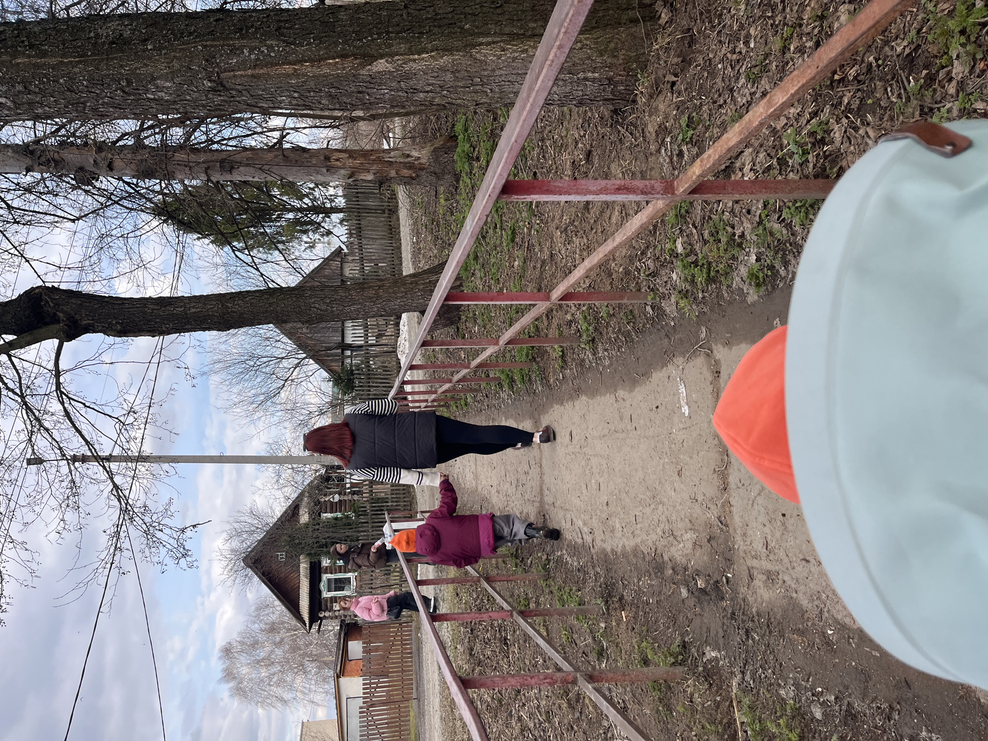 The image captures an outdoor scene, likely in a rural or semi-rural area of Chaikovsky, Russia, during what appears to be late winter or early spring, given the bare branches of the trees. The sky is bright with some clouds.
The primary subjects are several people walking along a dirt path. In the mid-ground, a woman with long red hair, wearing a black vest over a striped top and dark pants, walks away from the camera. Ahead of her is a small child in a purple jacket and an orange hat. Further down the path, another person in a brown jacket and a person in a pink jacket are also visible. In the very close foreground, parts of a light blue garment and an orange hat are seen, suggesting another person (possibly the photographer or a child) is standing near the camera.
A rusty red metal railing or fence runs along the right side of the dirt path. On the left side, several large trees with dark, textured bark and bare branches stand tall. In the background, traditional-looking wooden houses with dark roofs and some fences are visible, characteristic of village architecture. A utility pole with wires can be seen against the sky on the left. The ground is a mix of dirt, dry leaves, and patches of sparse green vegetation. The scene depicts a casual walk or outing for the group. There is no visible text in the image.