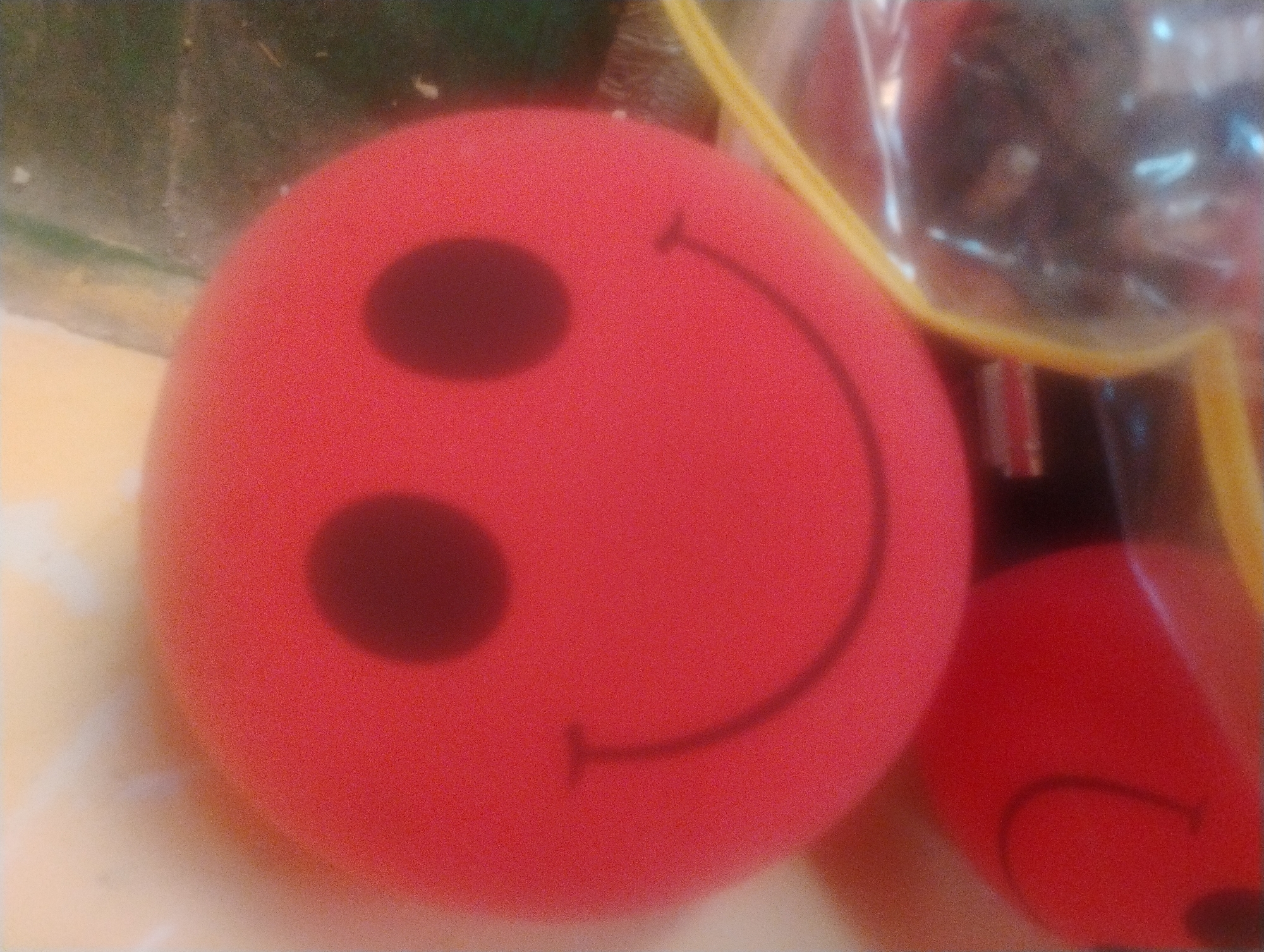A close-up, slightly out-of-focus image shows two red, spherical objects with smiley faces, possibly toy balls, against a blurred background. The object in the foreground is larger and displays a distinct smiley face with two dark, circular eyes and a curved mouth. A portion of a second, similar object is visible to the left. The objects are illuminated, creating soft shadows and highlights. The overall scene appears to be indoors, with hints of packaging and a textured surface beneath. There is no indication of the location being in Rumyantsevo, Bulgaria, or any other specific city.