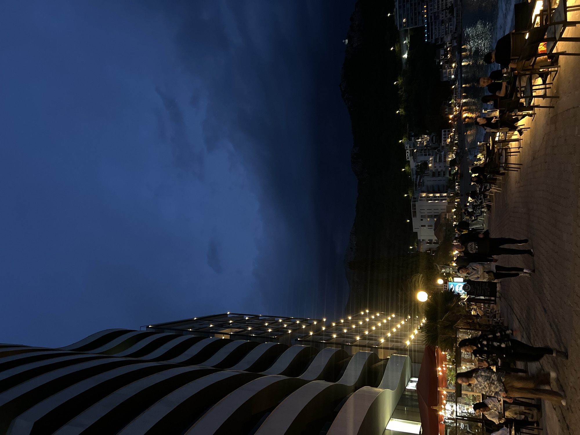 The image, taken at night, depicts a street scene in Rafailovići, Montenegro. The sky is dark blue and partly cloudy. A mountain range is visible in the background, dividing the sky. In the foreground, a modern building with a striped, wavy facade is illuminated by small lights. Along the right side of the image, people are walking along a brick-paved promenade. Tables and chairs are set up outside restaurants and cafes, occupied by patrons. The lights from buildings and streetlamps reflect on the water in the background.