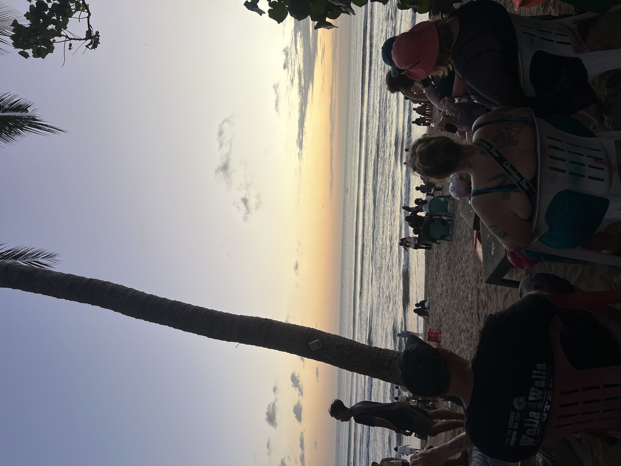 The image depicts a serene sunset scene on Kuta Beach, Indonesia. In the foreground, several people are seated on chairs on the sand, facing the ocean. A prominent palm tree trunk stands to the left, its fronds extending into the upper left corner of the frame. The ocean is calm with gentle waves rolling towards the shore. The sky exhibits a gradient of colors from pale blue at the top to warm orange and yellow hues near the horizon, indicating the sunset. Scattered clouds are visible in the sky.
In the middle ground, more people are scattered across the beach, some standing and others sitting on the sand. A few beach chairs and umbrellas are visible. The sea is populated with a few distant boats. The sand itself appears dark and wet closer to the water, suggesting the tide is out or receding.
The people in the foreground are mostly seen from behind, focusing on their relaxed postures as they observe the sunset. One individual in the front left is wearing a black t-shirt with visible text and graphics. To the right, two women are seated, one wearing a blue top with a designer strap and the other in a dark outfit with a red hat. In the middle ground, a young man is standing near the palm tree, looking out at the ocean. The overall atmosphere is one of tranquility and leisure, characteristic of a beach sunset.