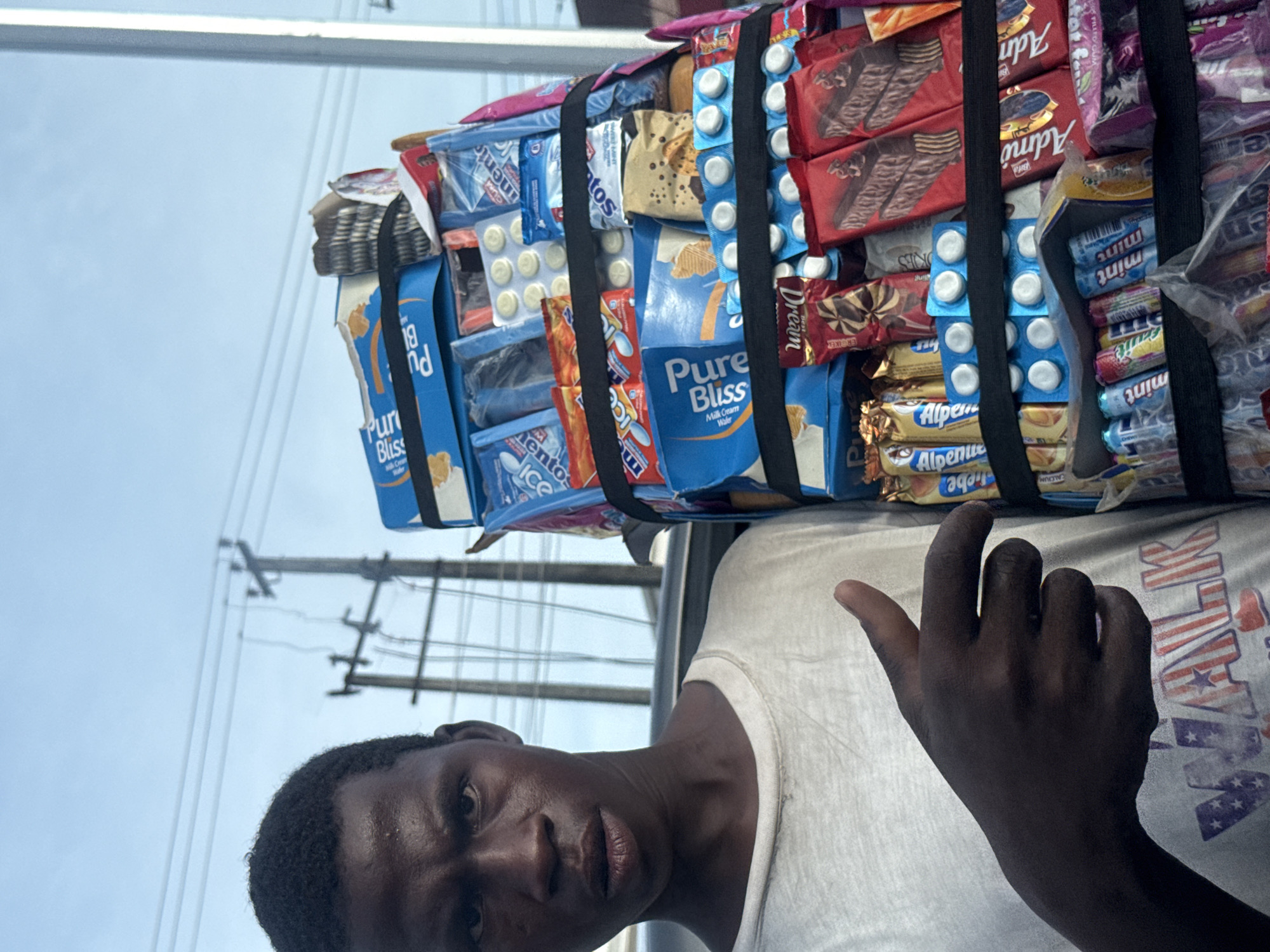 The image shows a dark-skinned man with short, dark hair looking towards the camera. He is wearing a white t-shirt with a faded American flag design on the right side. The man is carrying a large assortment of snacks and candies secured by black straps, including brands like "Pure Bliss," "Alpenue," "Admo," "Mentos," and "Dream," with blister packs of what appear to be tablets integrated within. The background shows a light-colored sky with electrical poles and wires visible. The context suggests this is taking place in Ado, Nigeria.