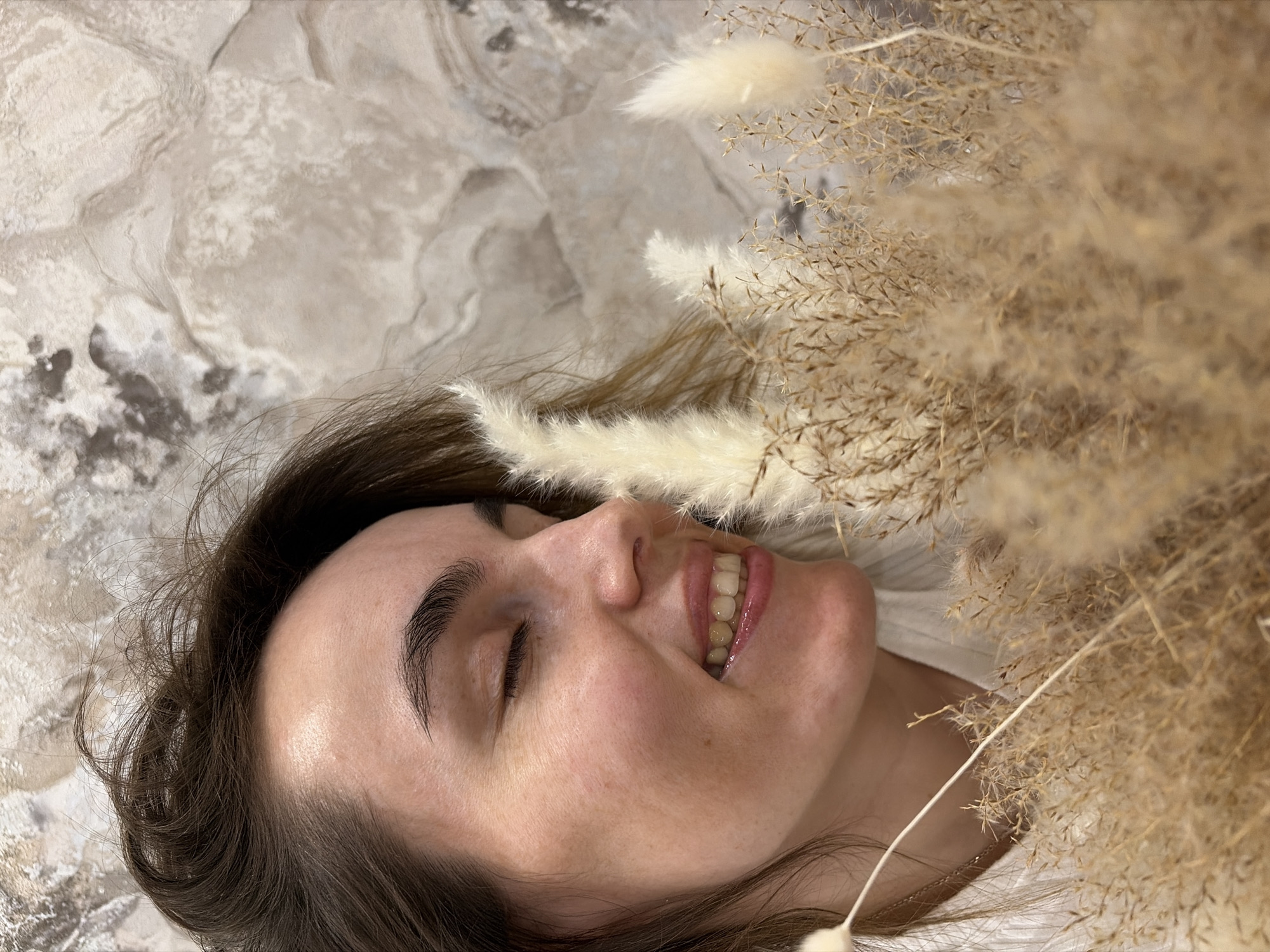 A woman is lying on a textured, marbled surface, with dried grasses arranged around her head. She has brown hair, closed eyes, and fair skin. She is wearing a white shirt. The background texture resembles a stone or marble pattern. The location is not discernible in the image.