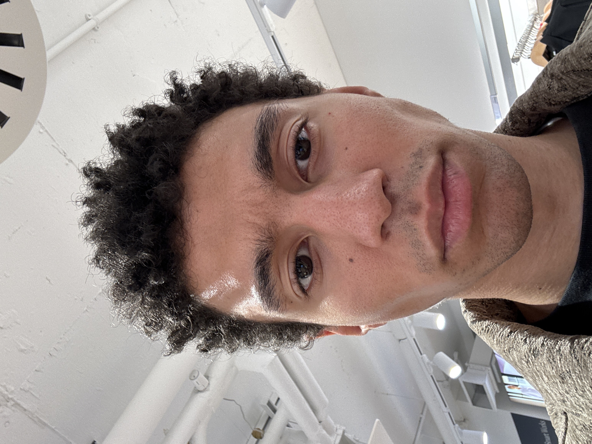 A close-up, slightly angled self-portrait of a man with tightly curled, dark hair and light brown skin. He has brown eyes and a light stubble. He is wearing a dark top with a textured brown jacket partially visible. The background is a bright, indoor space with white walls, ceiling fixtures, and some structural elements. The environment appears to be a store or commercial space due to the overhead lighting and display components in the background. There is no specific information available in the image to determine if it is located in London, United Kingdom.