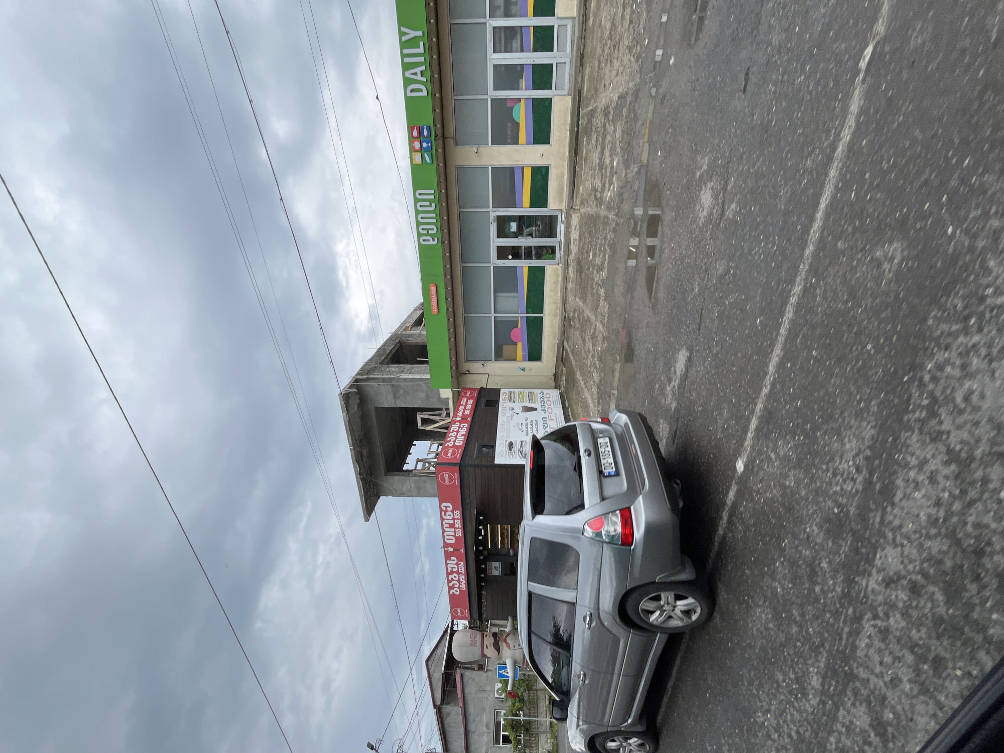 The image captures a street scene in Poti, Georgia, on a cloudy day. In the foreground, a silver Subaru Forester SUV is parked on the wet asphalt road. The road shows signs of recent rain with several puddles reflecting the overcast sky. To the right of the SUV, a modern building with a green awning and large glass windows is visible. The awning features colorful graphics and the word "DAILY" in large white letters. The building appears to be a commercial establishment, possibly a grocery store or convenience store. To the left of the "DAILY" building, another, older building is partially visible, with a red awning advertising food, featuring text in Georgian script and a phone number. Behind these buildings, a structure under construction is evident, with exposed concrete and scaffolding. The sky is heavily clouded, indicating a dull or rainy day. The overall scene depicts a typical urban street in Poti, with a mix of commercial activity and ongoing construction.