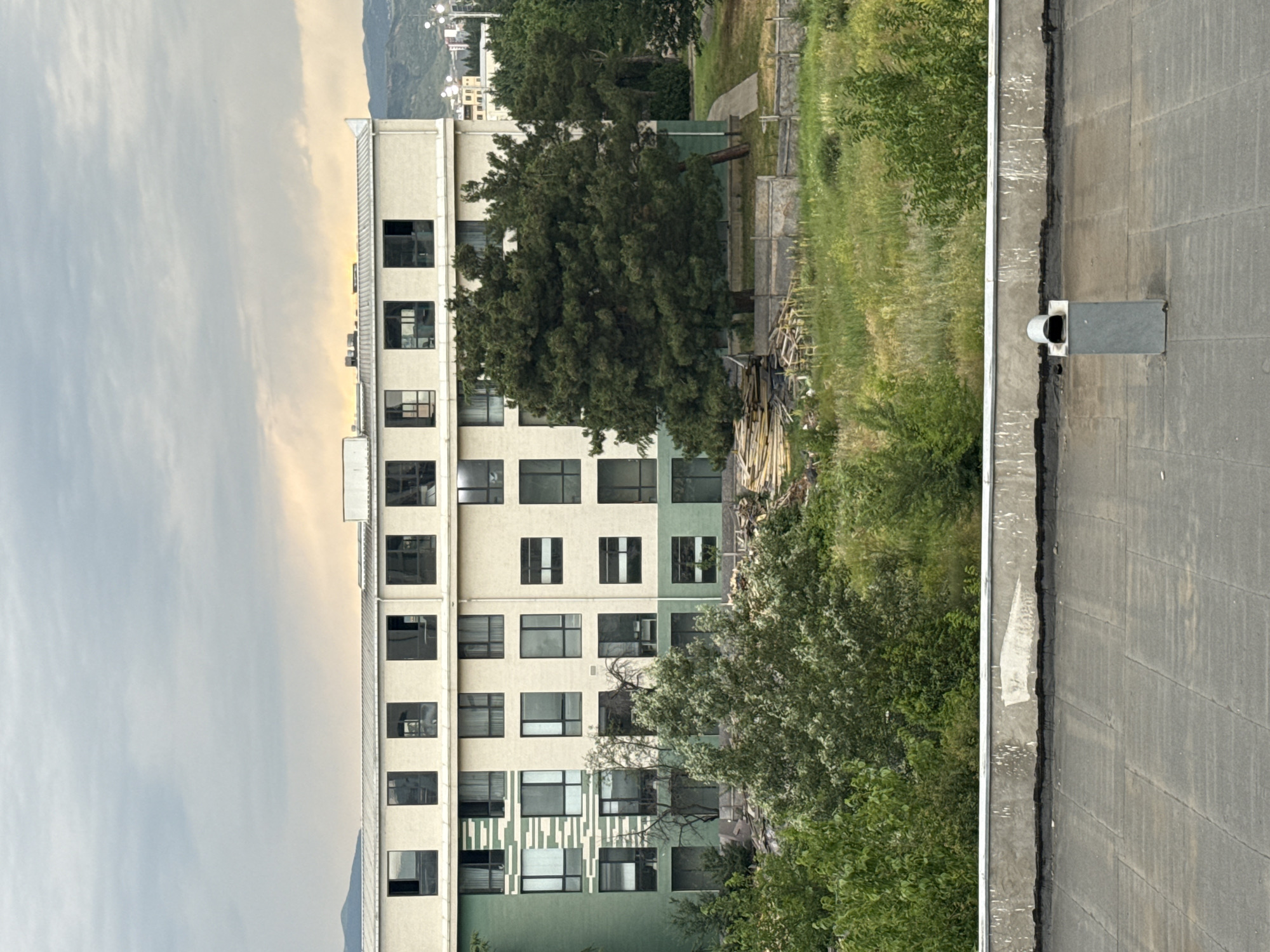 The image captures an overcast day in Tbilisi, Georgia, focusing on a multi-story building with a modern facade. The building features a symmetrical design with a light-colored upper section and a green lower section adorned with patterned windows. Large trees, characteristic of the region, surround the building, with a dense canopy of green foliage dominating the foreground. In the distance, rolling hills and urban structures suggest the presence of a city. The foreground shows a flat rooftop with a ventilation pipe, indicating an urban or semi-urban setting. The overall scene conveys a sense of residential or institutional architecture within a natural, verdant environment.