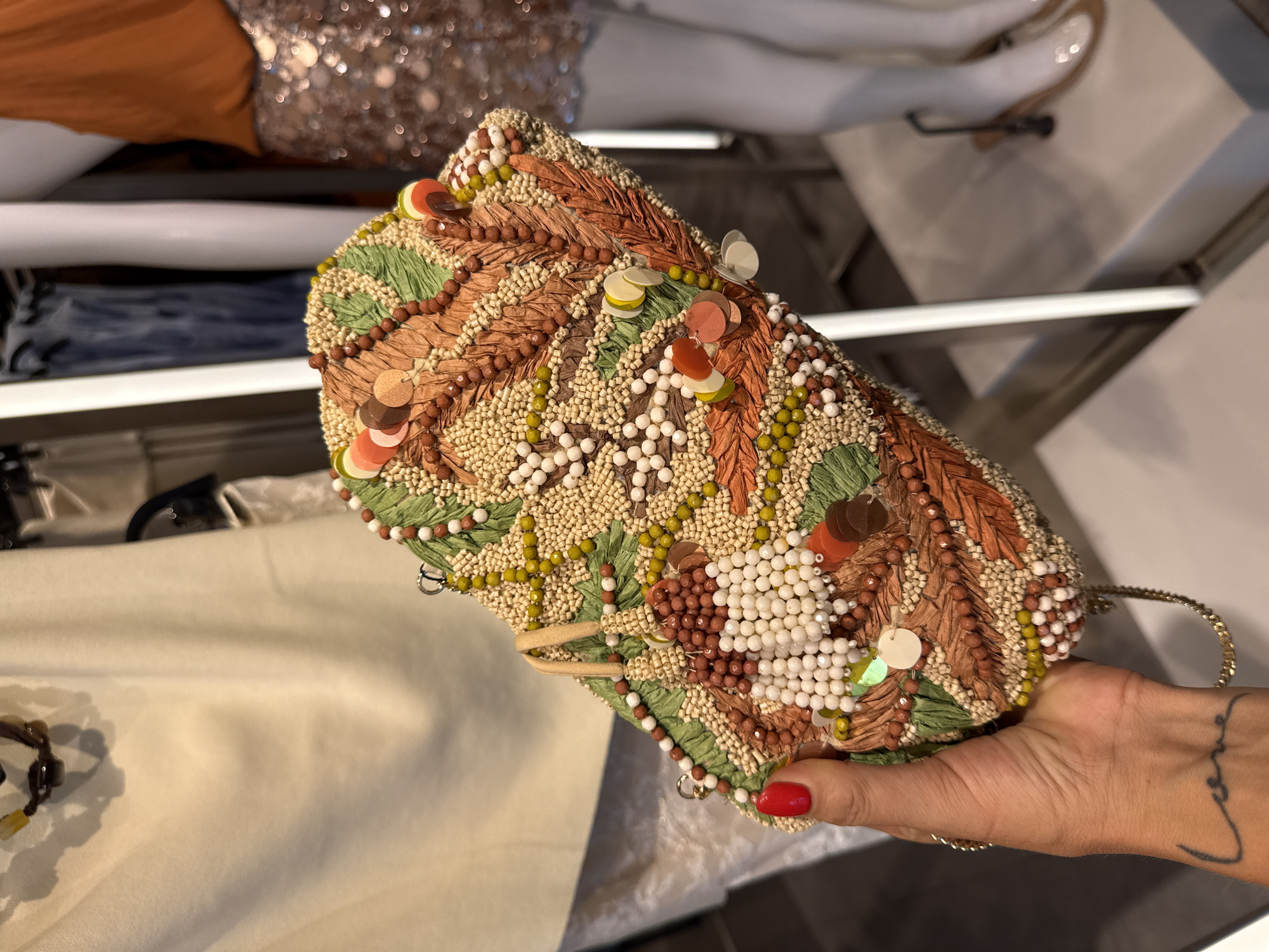 A beaded purse is held in a person's hand with red nail polish. The person has a tattoo on their wrist. The purse is brown, green, beige, and white, and decorated with beads, sequins, and raffia. The background contains clothing and shelves, suggesting the interior of a retail store. The location is likely Kuta, Indonesia.