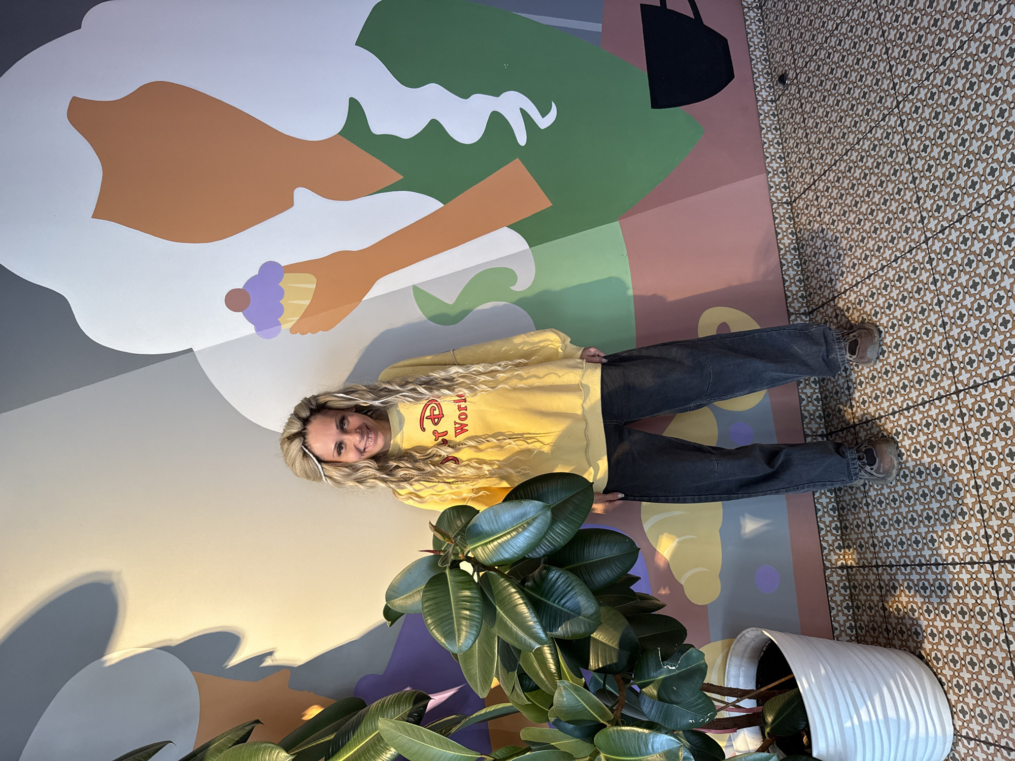 The image depicts a woman with long blonde hair standing indoors in front of a mural. She is wearing a yellow sweatshirt with "Disney World" written on it and dark jeans. To her right is a large potted plant with glossy green leaves. The mural features stylized figures and shapes in various colors including white, gray, brown, green, pink, purple, and yellow. The floor is tiled with a geometric pattern. The exact location is Rusanovo, Russia.