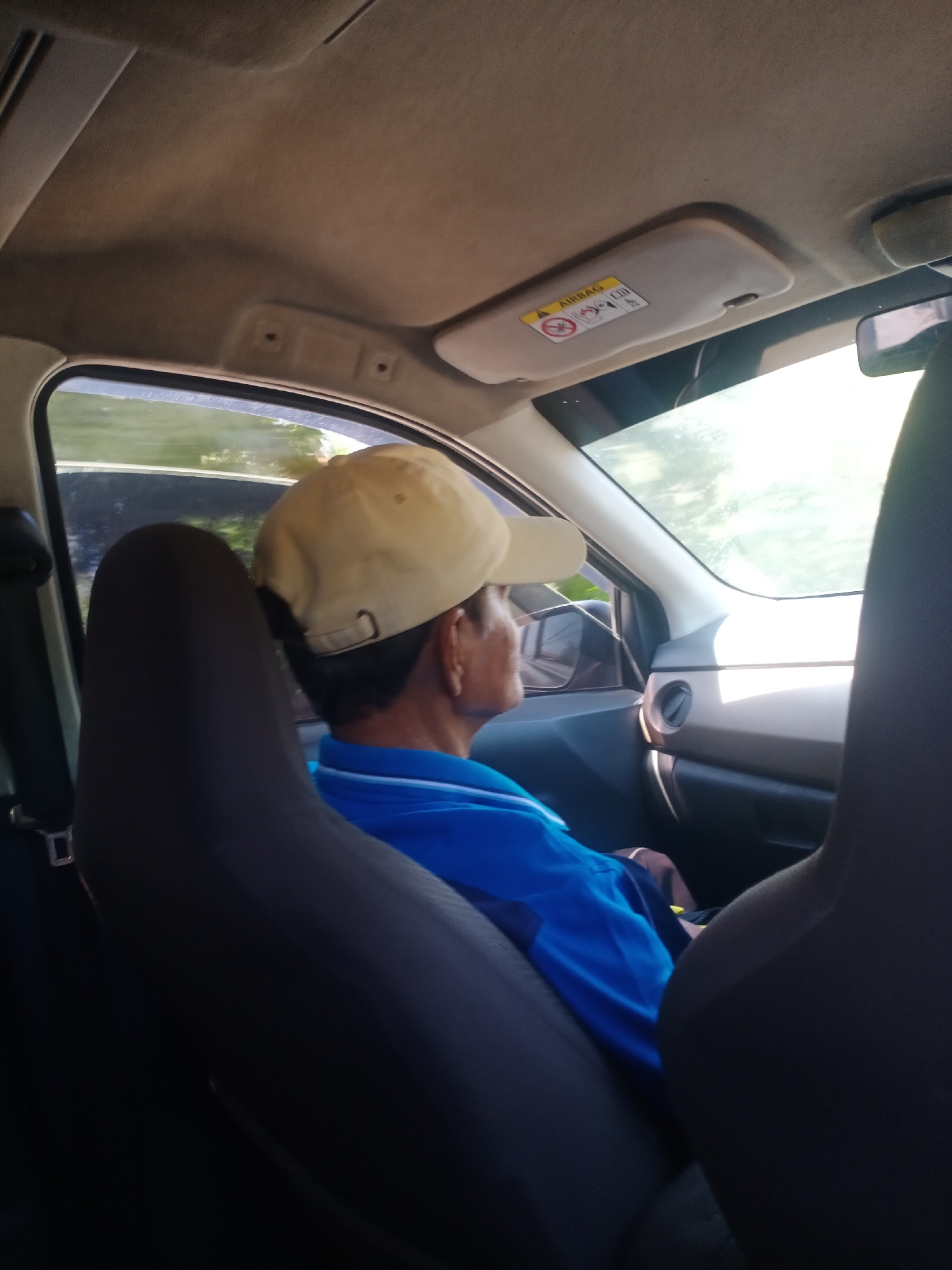 The image shows the interior of a car with a person sitting in the passenger seat. The person is wearing a light-colored baseball cap and a blue shirt. The view is from behind the passenger, looking towards the front of the car and out the side window. The sun is shining brightly through the window, creating a glare. The dashboard and part of the driver's seat are visible. There is an "AIRBAG" warning label on the sun visor. The context of Lalolang, Indonesia, is not discernable from the image itself.