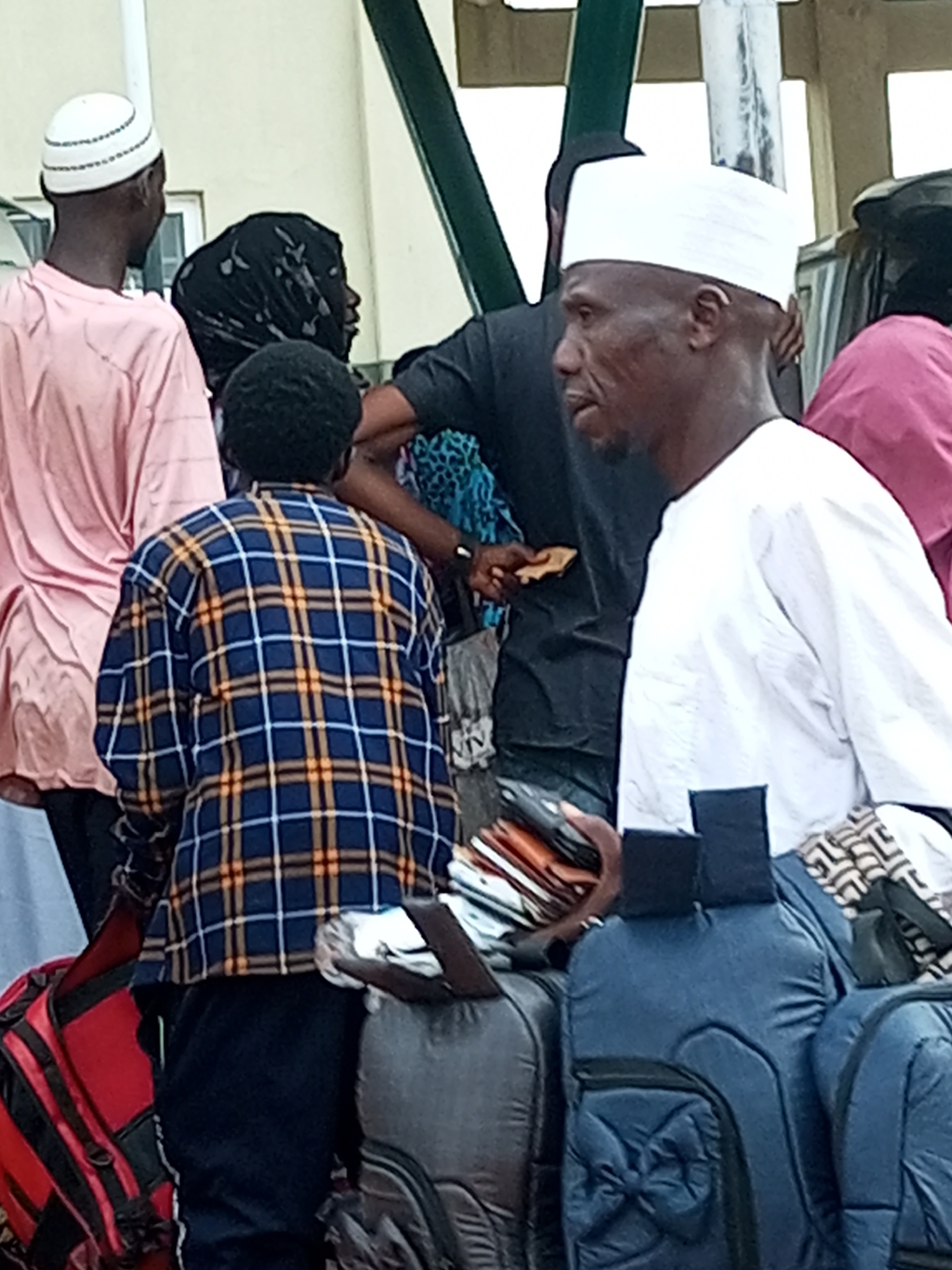 In Gombe, Nigeria, a group of people are gathered near a covered structure, possibly a bus station or shelter. The scene appears to be outdoors with a light-colored wall and green supporting poles visible in the background.
In the foreground, a man wearing a white kufi and a white long-sleeved garment faces to the left, with a slight smirk. He is carrying a blue duffel bag and appears to be interacting with someone or observing the surroundings. To his left, another man, also wearing a white kufi, is seen from the back, dressed in a pink long-sleeved shirt. He is looking away from the camera.
Behind these two individuals, a person in a black hijab is partially visible. In front of the person in the pink shirt and to the right of the man in the white garment, a person wearing a blue and yellow plaid shirt is also seen from the back, with their arms down. Several pieces of luggage are present, including red and black backpacks, a gray duffel bag, and multiple dark-colored bags, some stacked. The overall impression is one of waiting or transit.
