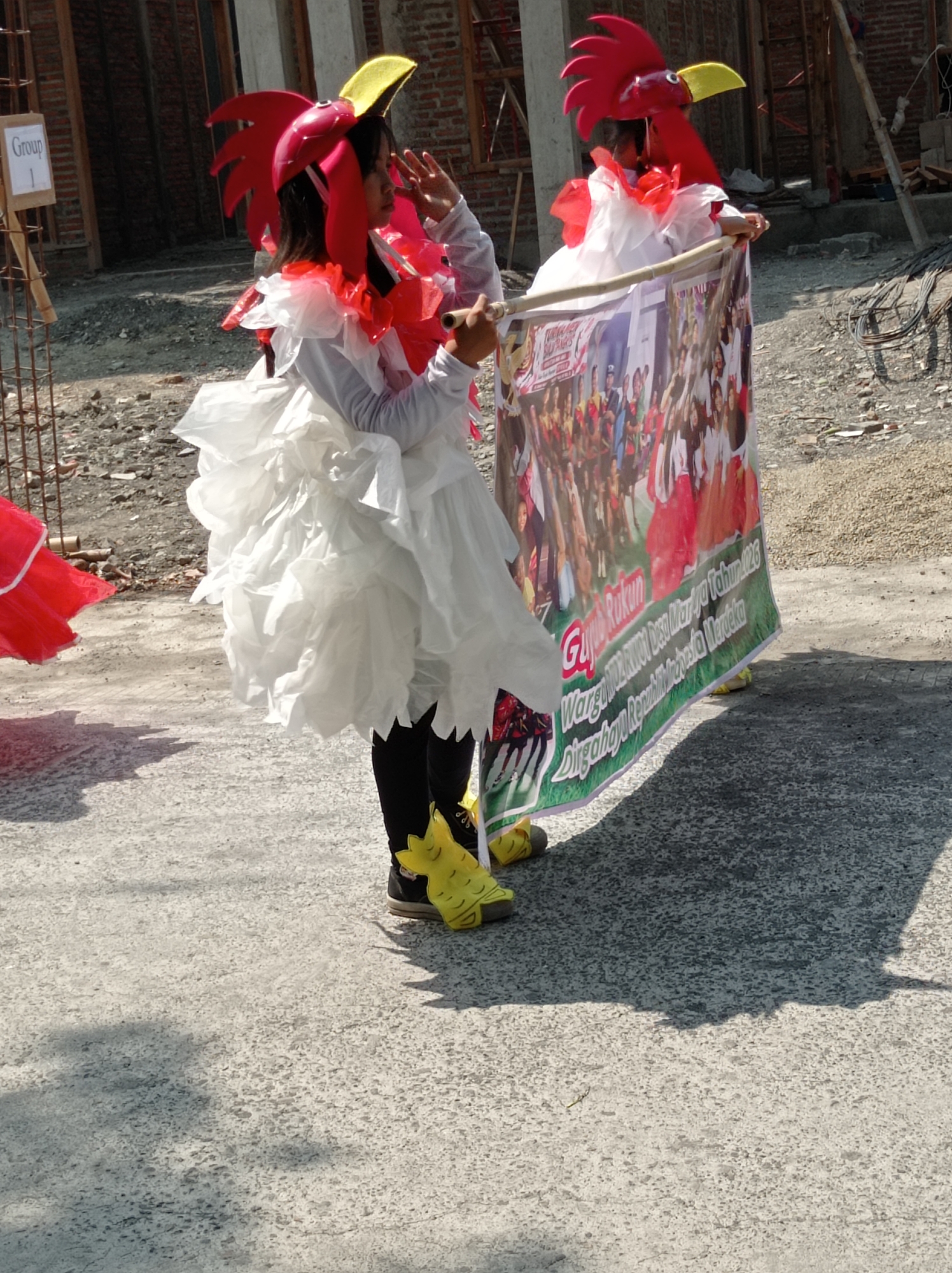 Two young girls, dressed in elaborate rooster costumes with red and yellow headdresses and white ruffled outfits, participate in a parade in Maribaya, Indonesia. One girl holds a banner with a photograph of many people and Indonesian text that reads "Gajah Rukun Warga RW 01 Desa Maribaya Tahun 2023 Dirgahayu Republik Indonesia Merdeka". The other girl stands beside her, holding a pole. Both girls wear yellow shoe covers. The ground is paved with asphalt. In the background, construction scaffolding and brick walls are visible, suggesting an area undergoing development. A sign with "Group 1" written on it is attached to a wooden post.