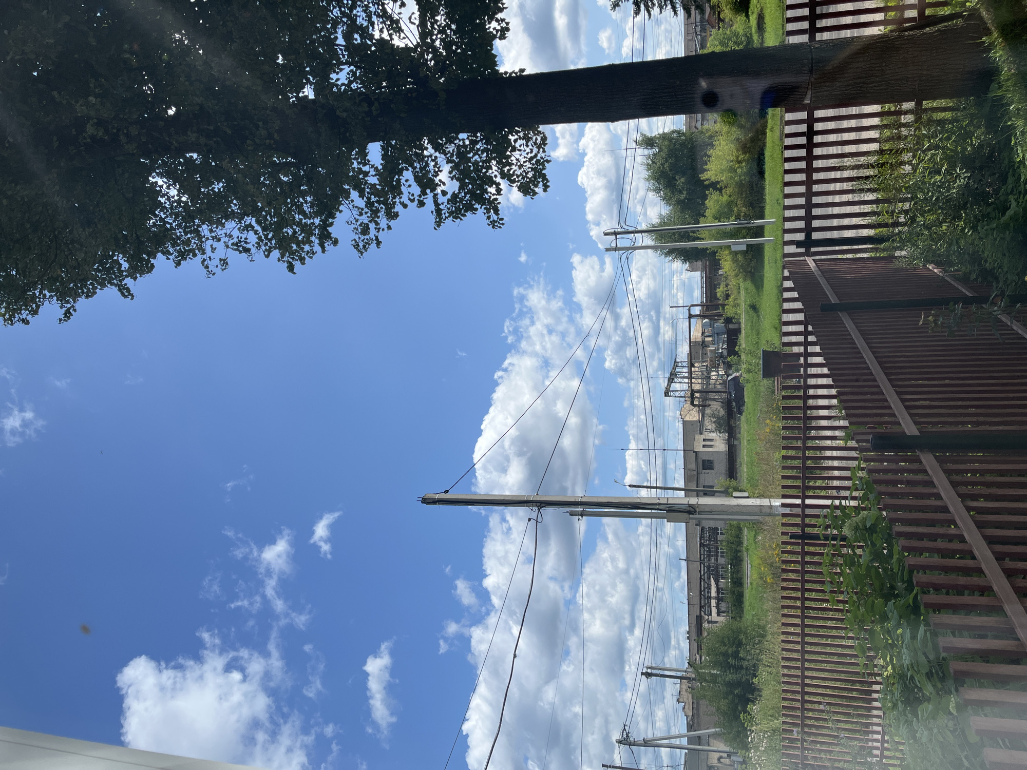 This image shows a view of an industrial area in Ivanovo, Russia, on a sunny day with a blue sky and scattered clouds. In the foreground, there is a wooden fence with green vegetation behind it. A tall tree with dark green leaves is on the right side of the image, partially obscuring the sky. Beyond the fence and vegetation, there are several power lines and utility poles stretching across the scene. In the background, a row of industrial buildings and structures can be seen, surrounded by greenery. The overall impression is one of an urban fringe with industrial activity.