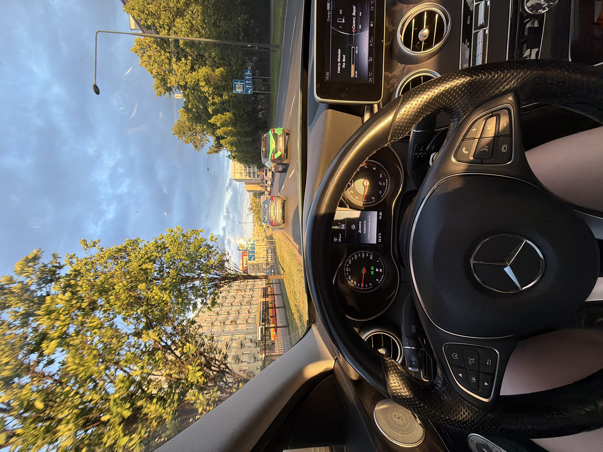 A Mercedes-Benz interior view of the road ahead in the city of Unknown, Unknown. The dashboard display shows the music artist Valeriy Meladze's song "The Best" playing and a temperature of 24°C. The speedometer registers 34 km/h. Outside, traffic is moving down a city street lined with trees and buildings.