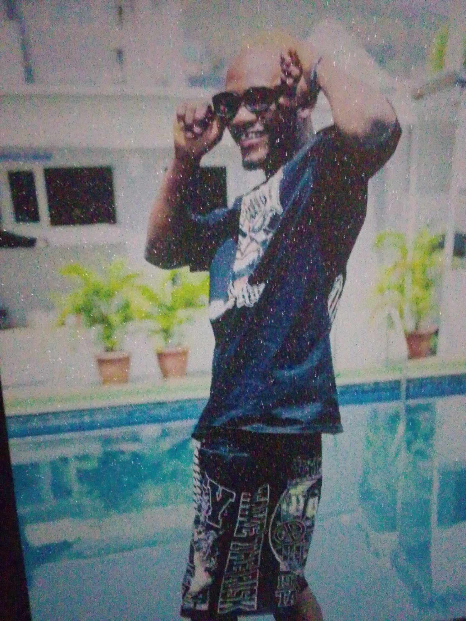 A bald, dark-skinned male with sunglasses is posing next to a swimming pool. He is wearing a dark t-shirt and patterned shorts. In the background, there are potted plants and a building. The image is blurry and grainy, making it difficult to discern fine details. The scene appears to be outdoors, possibly at a private residence or a recreational facility. There is no indication of the specific location in Keffi, Nigeria.