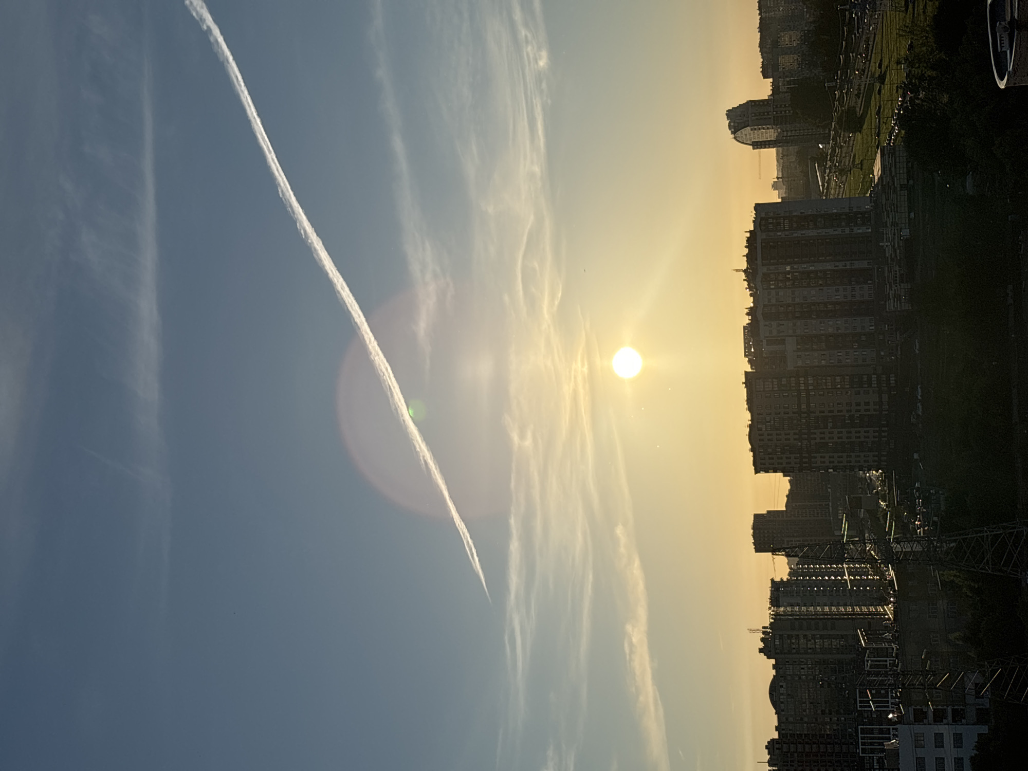 The image depicts a cityscape during what appears to be sunset. The sky is a gradient of light blue and yellow, with thin, wispy clouds. A prominent contrail from an airplane stretches diagonally across the upper portion of the sky. The sun, a bright orb, is visible in the middle of the frame, casting a warm glow. Below the sky, the city skyline is dominated by numerous tall residential buildings of varying architectural styles and heights, densely packed together. The buildings are silhouetted against the bright sky, with some windows reflecting the sunset light. In the foreground, industrial structures, possibly power lines or towers, are visible, along with dark trees and foliage. The scene captures an urban environment at the end of the day, with the sun setting over the buildings. The location is identified as unknown, Russia.