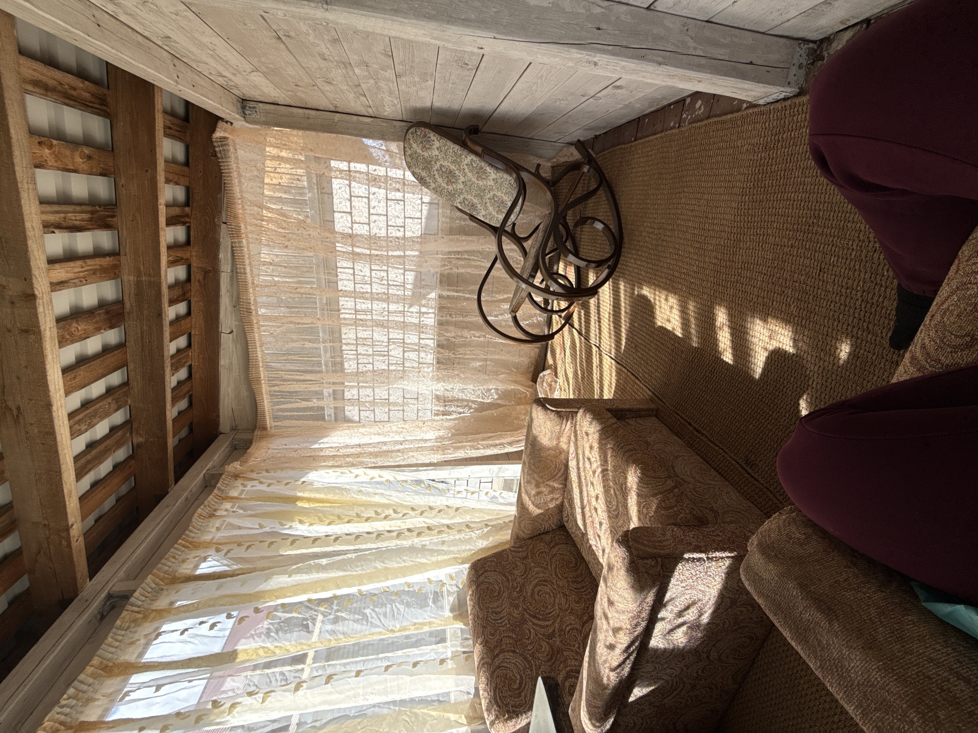 An interior space is illuminated by sunlight filtering through a window covered with sheer curtains. A rocking chair with a floral-patterned seat sits near the window. A brown rug covers the floor. A person wearing burgundy pants is seated on an armchair. The ceiling is made of wooden planks supported by wooden beams. The location is Yakshur-Bod'ya, Russia.