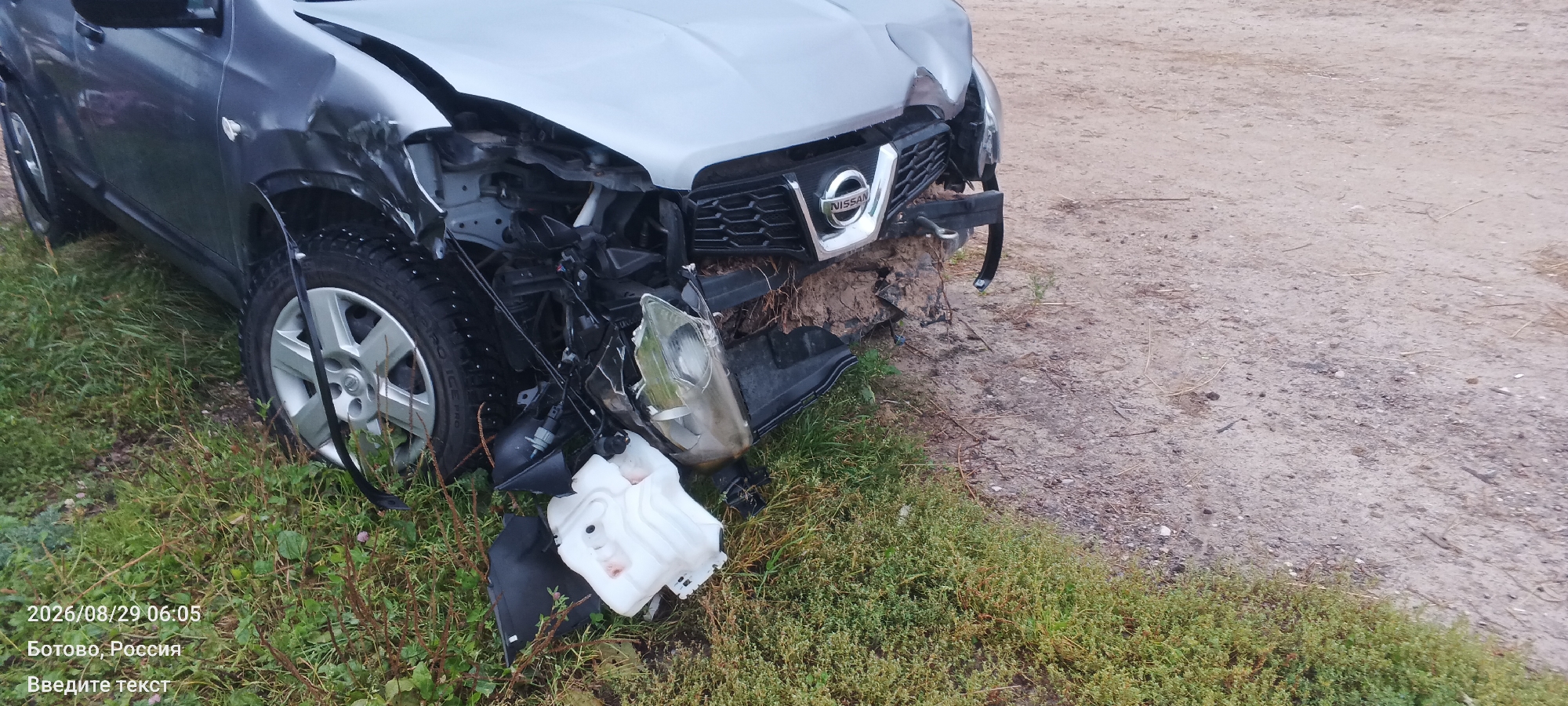 A damaged silver Nissan Qashqai is parked on the side of a dirt road, with its front end heavily impacted. The bumper is detached, the hood is dented, and the headlight is broken. The front wheel on the driver's side is turned outwards, and some engine components are exposed. The car is surrounded by green grass and a dirt path. The timestamp 2026/08/29 06:05 and the text "Ботово, Россия" (Botovo, Russia) and "Введите текст" (Enter text) are overlaid on the bottom left of the image.