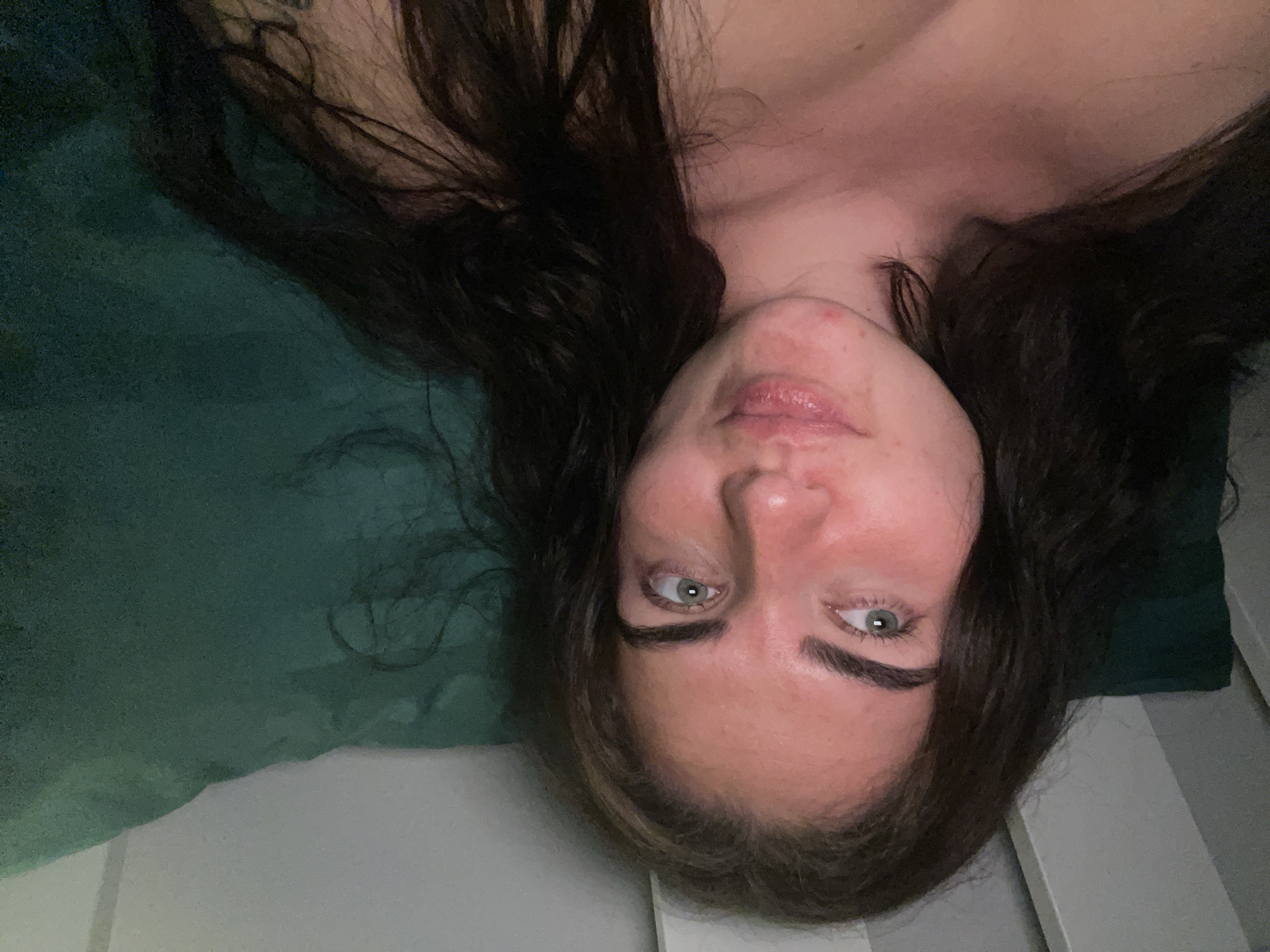 The image shows a close-up, waist-up view of a young woman lying down on a dark green surface, likely a pillow or couch. She has long, dark brown hair that falls around her shoulders and a fair complexion with some redness on her cheeks and chin. Her eyes are a light green and are looking off to the side. She is wearing a low-cut garment that exposes her upper chest. The lighting is dim, with a pinkish hue illuminating her skin. The background consists of a white vertical surface on the left and the dark green surface she is lying on. There is no discernible indication of her location within the city of Мосметаллоконструкция Онт, Russia.