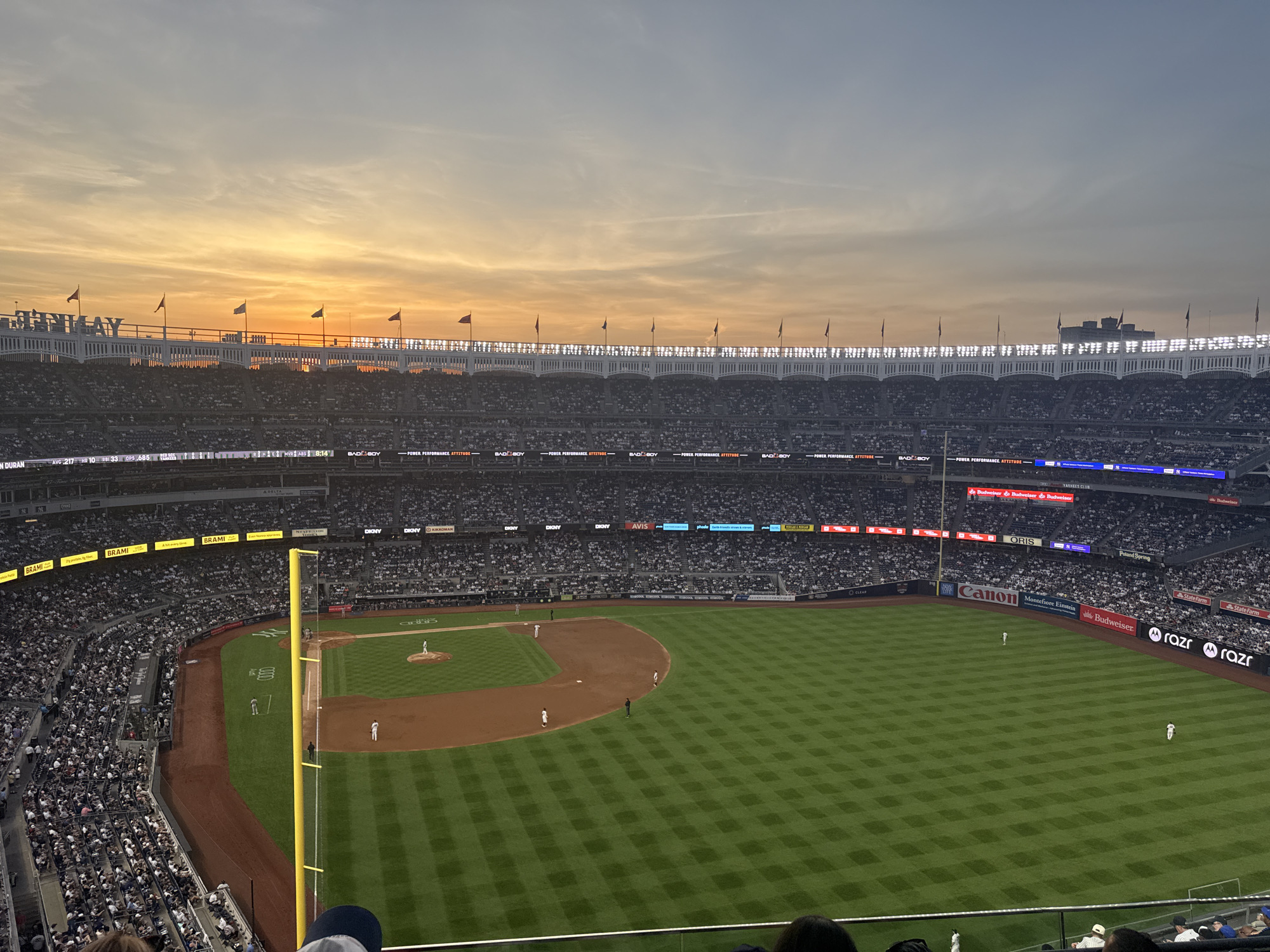 This is an elevated, wide-angle photograph of an evening baseball game in progress at Yankee Stadium, located in The Bronx, New York City, United States of America. The sky above the stadium displays a sunset with vibrant orange and yellow hues transitioning into a pale blue-grey. Stadium lights are illuminated on the upper deck.
The baseball field, featuring a checkerboard pattern mown into the green grass outfield and a reddish-brown dirt infield, occupies the lower and central portions of the frame. Several baseball players, distinguishable by their white uniforms, are visible on the field: one on the pitcher's mound, one near first base, and at least two outfielders. A tall yellow foul pole stands prominently on the left side of the field.
The stadium stands are densely populated with spectators, appearing as small white and dark figures across multiple tiers. The stadium structure includes white railings and advertising hoardings displaying various sponsor logos such as "Yankees," "BRAMI," "DKNY," "KIKKOMAN," "AVIS," "Canon," "Budweiser," "razr," "State Farm," "ORIS," "Poland Spring," "CLEAR," and "Montefiore Einstein." A digital clock displaying "8:14" is also visible. Numerous small flags, primarily red and green, fly atop the upper deck railing against the sunset sky. The perspective is from an elevated seat within the stadium, looking towards the field and the opposing stands.