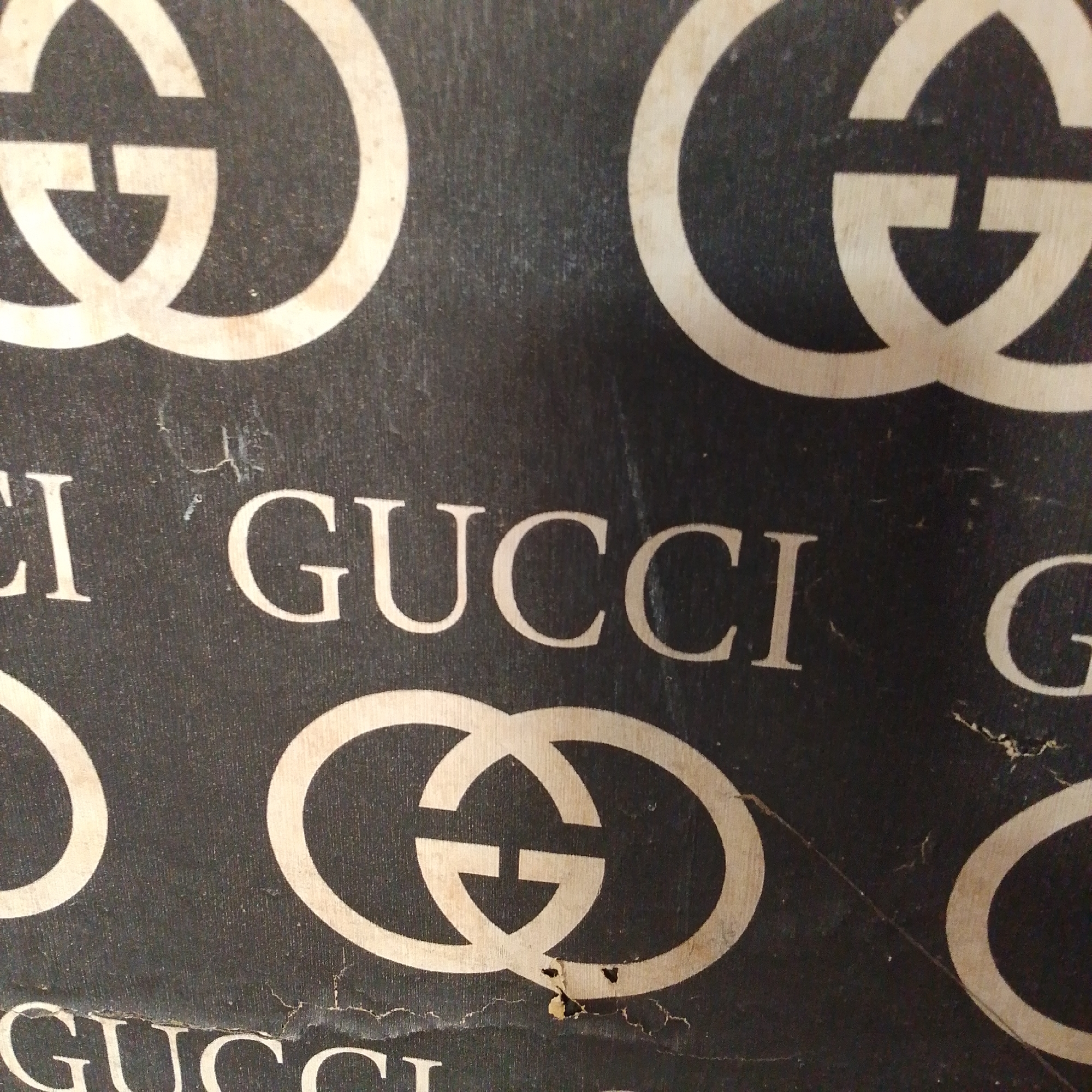 The image displays a close-up view of a black surface with a repeating pattern of light-colored Gucci logos and the word "GUCCI" printed in capital letters. The logos are the interlocking Gs characteristic of the brand. The word "GUCCI" appears twice, with one instance partially visible on the left side of the image. The texture of the black surface appears somewhat rough, with visible fibers and some signs of wear, including small tears and scuff marks. The lighting is diffused, casting subtle shadows and highlighting the texture. There are no people or any other identifiable objects or context in the image beyond the brand elements and the textured surface. The location is not discernible from the image content.