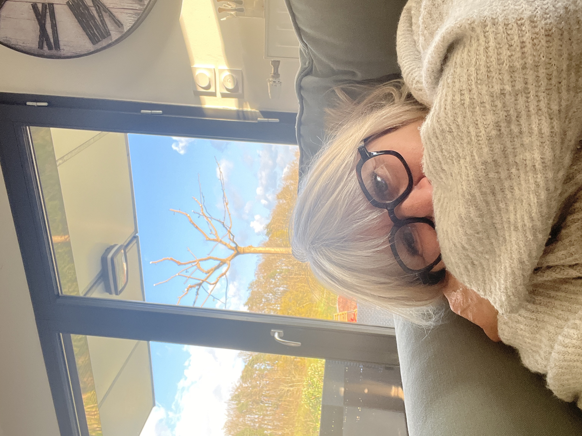 A middle-aged woman with short, gray hair and black-rimmed glasses is lying on a green sofa, her face partly covered by a beige knitted sweater. A window behind her shows a tree without leaves and a backdrop of trees with yellow and green foliage against a partly cloudy blue sky. A clock with Roman numerals hangs on the wall above the window. The location is in Chambœuf, France.