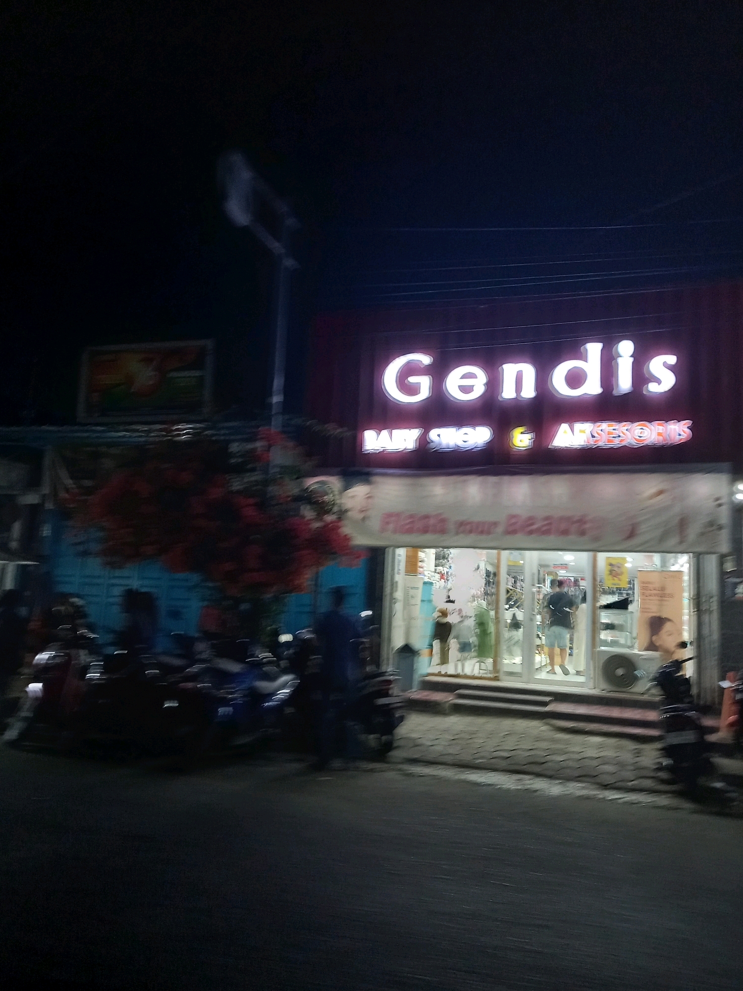 The image shows a storefront at night in Bawang, Indonesia. The storefront has a large illuminated sign that reads "Gendis BABY SHOP & AKSESORIS." Inside the store, there are displays of merchandise, and a person is visible behind the counter. In front of the store, there are several parked motorcycles. The street in front of the store is paved, and there are some trees with red flowers. The overall scene suggests a commercial area that is active at night.