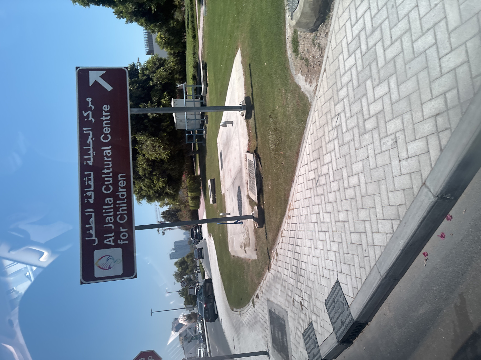 A brown sign with white text and a white arrow pointing to the right indicates "Al Jalila Cultural Centre for Children". The sign also has an Arabic inscription above the English text. The sign is attached to a metal pole on the right side of the image. To the left of the sign, there is a sidewalk made of light gray pavers, a street with gray asphalt, and a green grassy area. Several cars are visible on the street in the distance, and some buildings and trees are also present in the background under a clear blue sky. The scene appears to be in an urban or suburban environment in Dubai, United Arab Emirates.