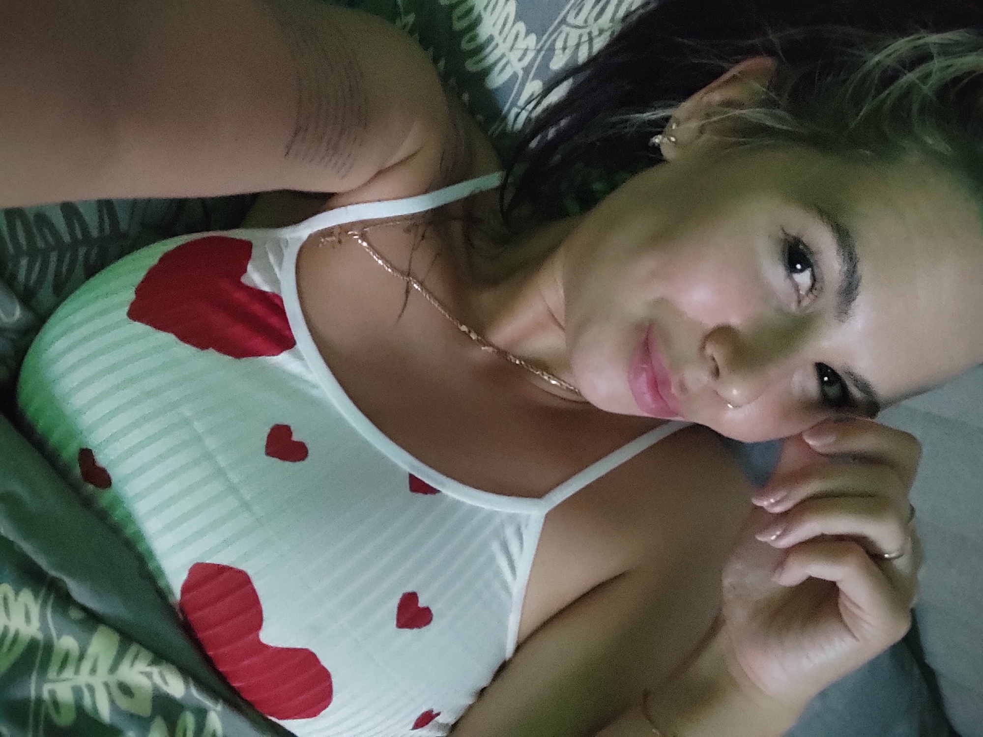 A woman lies on a bed in Chaikovsky, Russia. She wears a white camisole top with red hearts, and a gold necklace. Her dark hair is pushed to the side. Her right hand is near her face. The bedding is patterned with leaves and gray shapes.