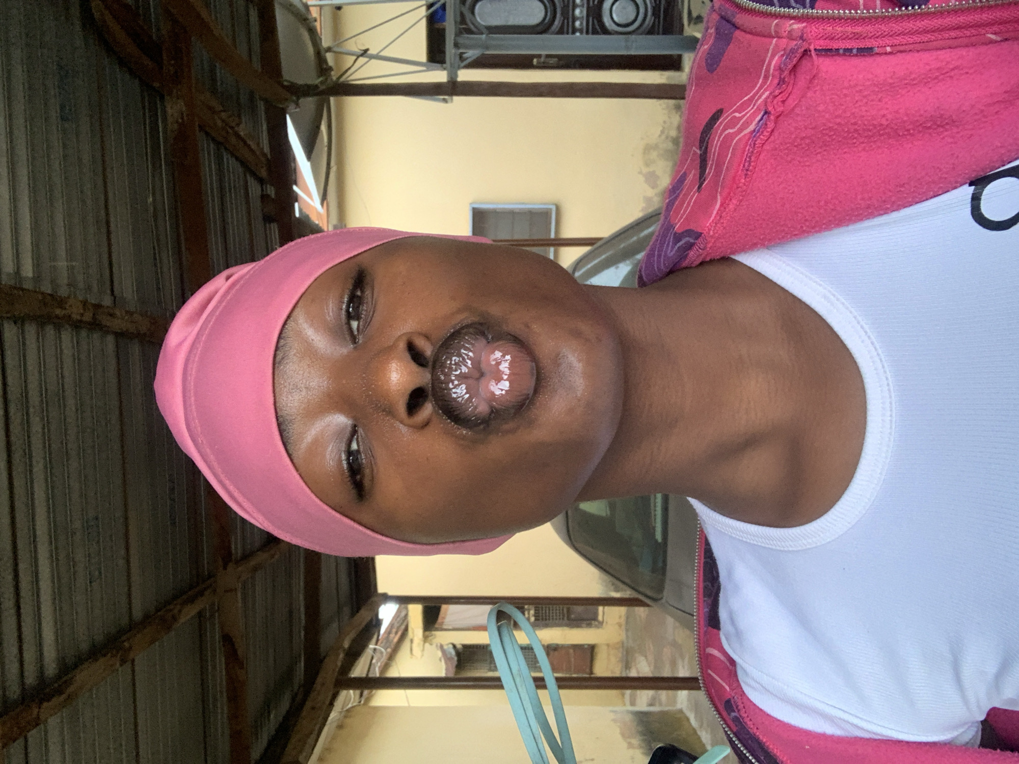 A woman with dark skin is posing for a selfie. She is wearing a pink headscarf, a white tank top, and a pink jacket. She is pouting her lips. The background includes a building with a yellow exterior, a car, and a metal structure. The location is Rafin Par, Nigeria.