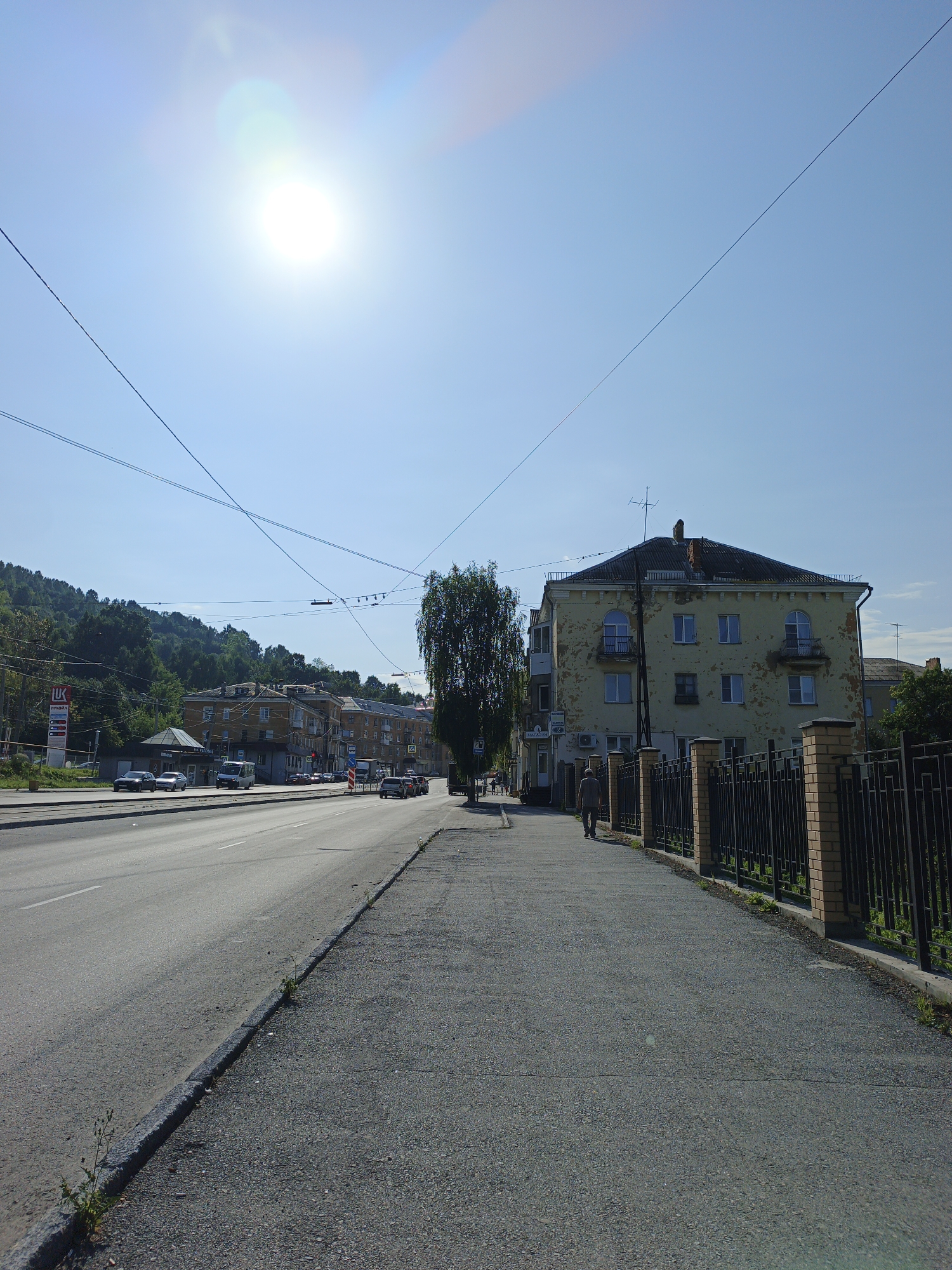 The image depicts a street scene in Zlatoust, Russia, under a bright, sunny sky with visible lens flare from the sun. In the foreground, a paved sidewalk runs along the right side of the frame, bordered by a black metal fence with brick pillars. A person is walking away from the camera on the sidewalk. To the left of the sidewalk is a multi-lane asphalt road with vehicles in motion.
Across the road, several buildings are visible. The most prominent is a yellow, multi-story building with a dilapidated facade, featuring arched windows and balconies. Further down the street, other residential buildings and some commercial establishments, including a gas station with prominent signage, are present. In the background, a lush green, tree-covered hill rises, suggesting a proximity to nature within the urban environment. Overhead, numerous power lines crisscross the sky, a common sight in Russian cities. The overall impression is of a typical urban street on a clear, warm day.