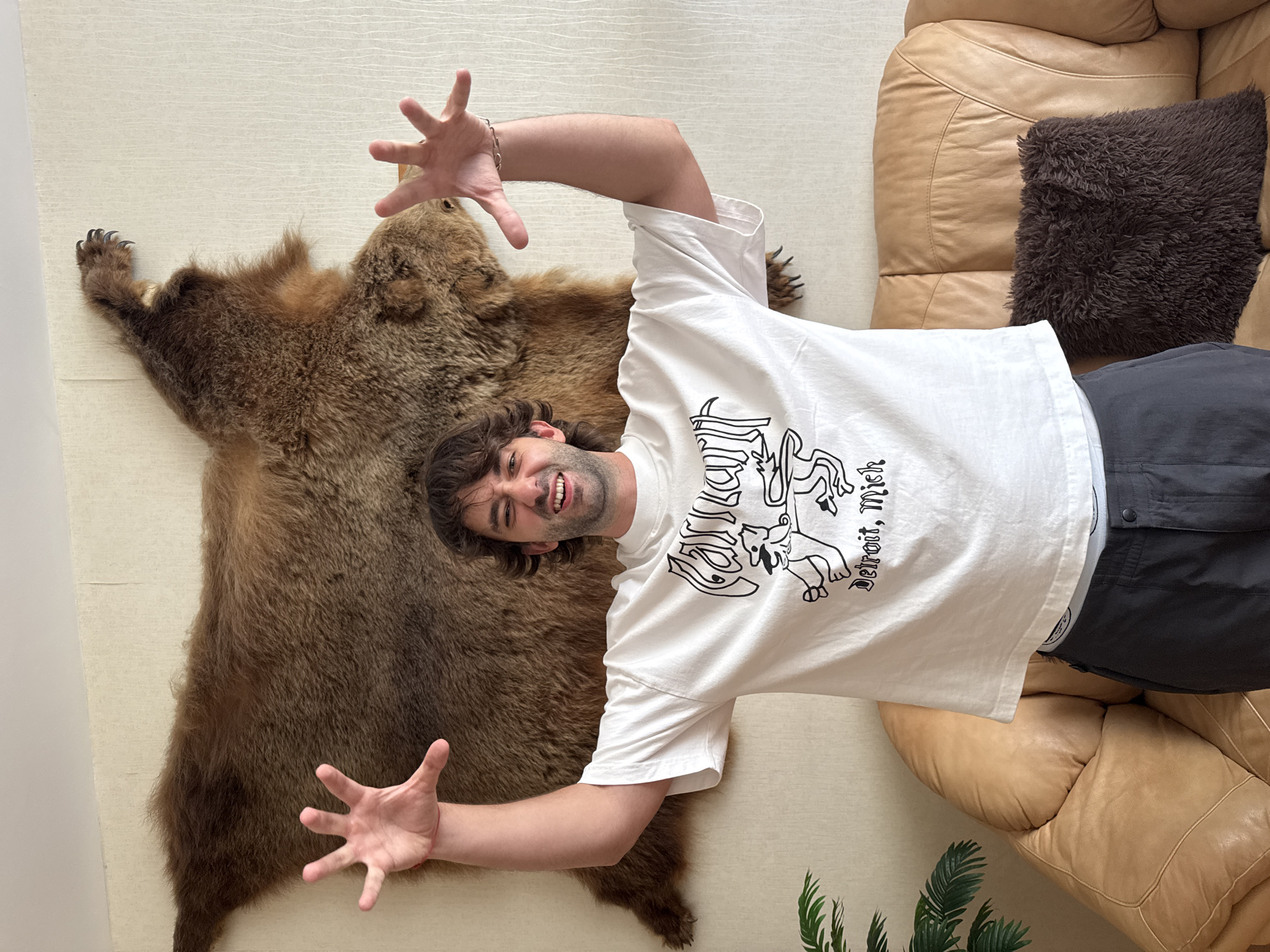 A man is lying on a light-colored textured surface, on top of a large brown fur rug that appears to be from a bear. The man has dark, curly hair and is smiling with his mouth open, looking towards the camera. He is wearing a white t-shirt with a black graphic print and dark gray pants. His arms are outstretched, with his left arm resting on the rug and his right arm reaching upwards. His hands are open with fingers spread. To the right of the man is a large, plush, tan leather sofa with a dark brown furry cushion on it. The scene appears to be indoors, with a neutral-colored wall on the left. There is visible text on the man's t-shirt that reads "Detroit, Mich." The overall impression is that of a playful or candid moment captured indoors. The image context suggests this is in Nizhny Novgorod, Russia, although there are no specific visual cues in the image to confirm this location. There are no indicators of time of day or weather conditions.