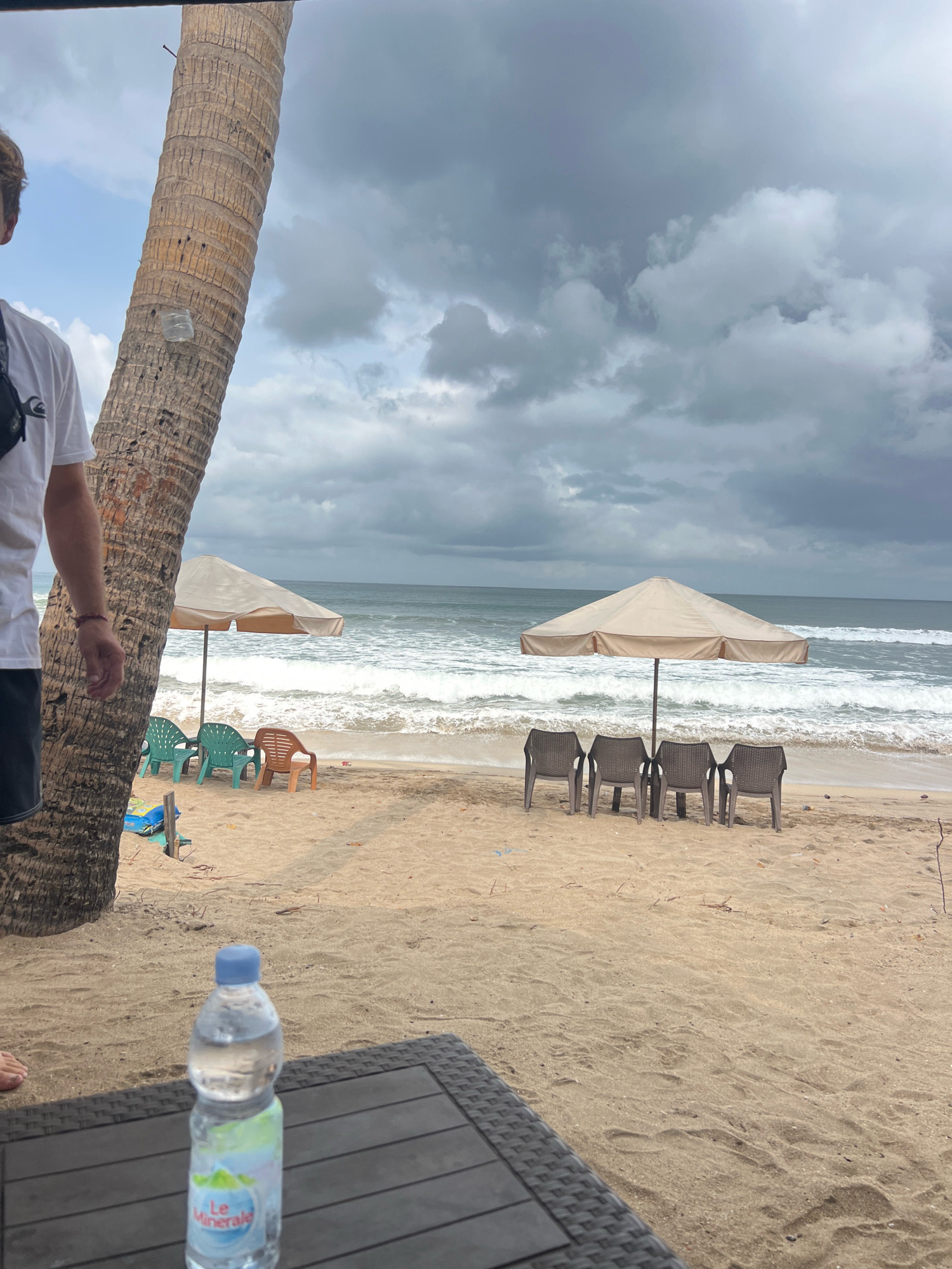 A beach scene in Kuta, Indonesia, features a cloudy sky over the ocean with moderate waves. In the foreground, a person in a white t-shirt and dark shorts stands partially obscured by a large palm tree on the left. A table with a plastic bottle of water is positioned in the lower foreground. The sand on the beach is light brown. Several beach chairs are arranged in two groups under two large, light-colored umbrellas. One umbrella has three green chairs and one brown chair in front of it, while the other umbrella has four dark-colored chairs lined up. The ocean extends to the horizon, with white foam visible from the breaking waves.