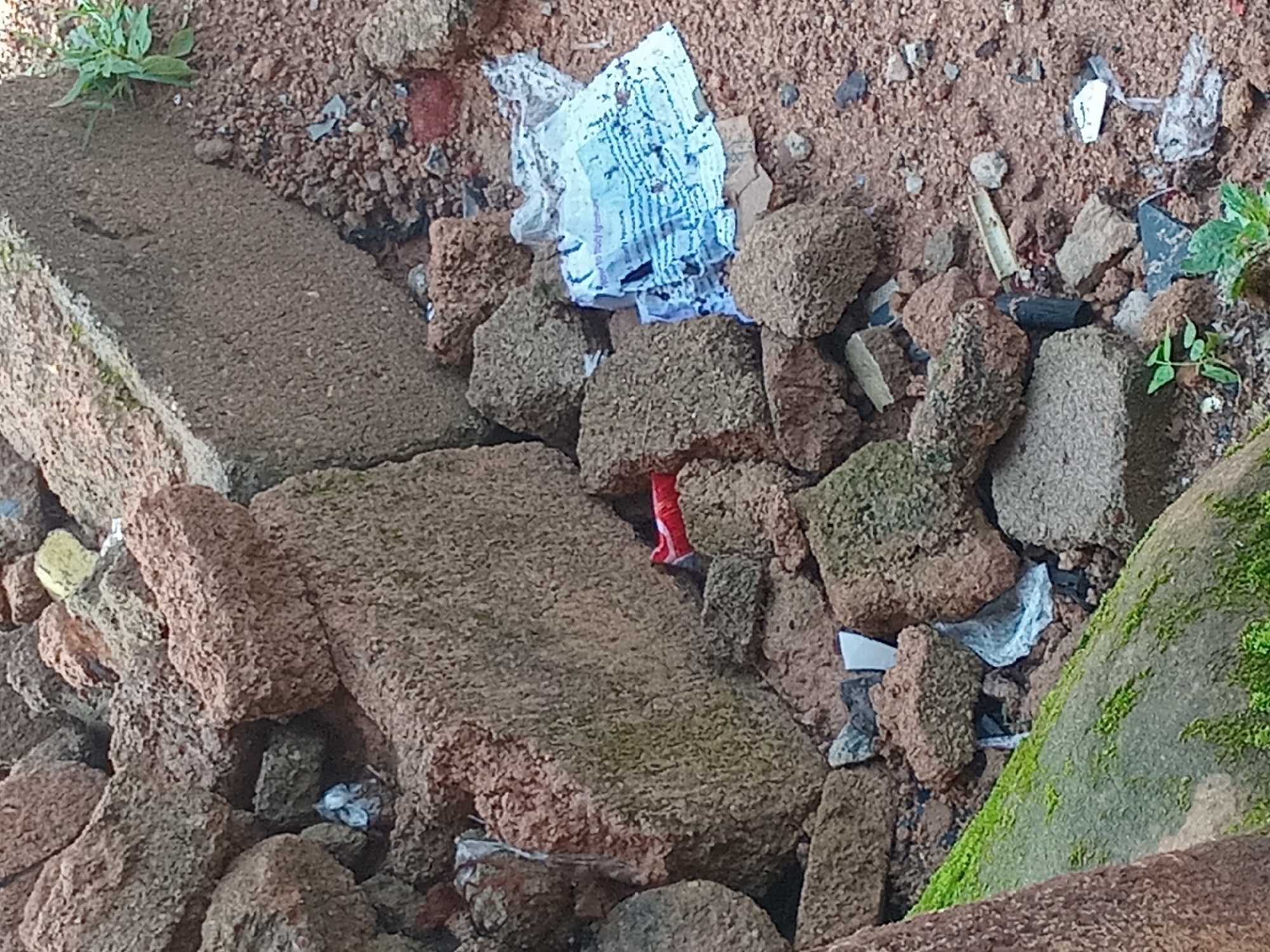 The image depicts a pile of rubble, primarily consisting of broken bricks and concrete blocks. Among the debris, there is some scattered trash, including crumpled paper and plastic. The scene is set outdoors, with visible soil and small green plants growing among the debris. The ground is uneven and covered in dirt and smaller stones. In the distance, a larger concrete structure is partially visible. The lighting suggests it is daytime, with natural light illuminating the scene. The overall impression is one of disarray and neglect.