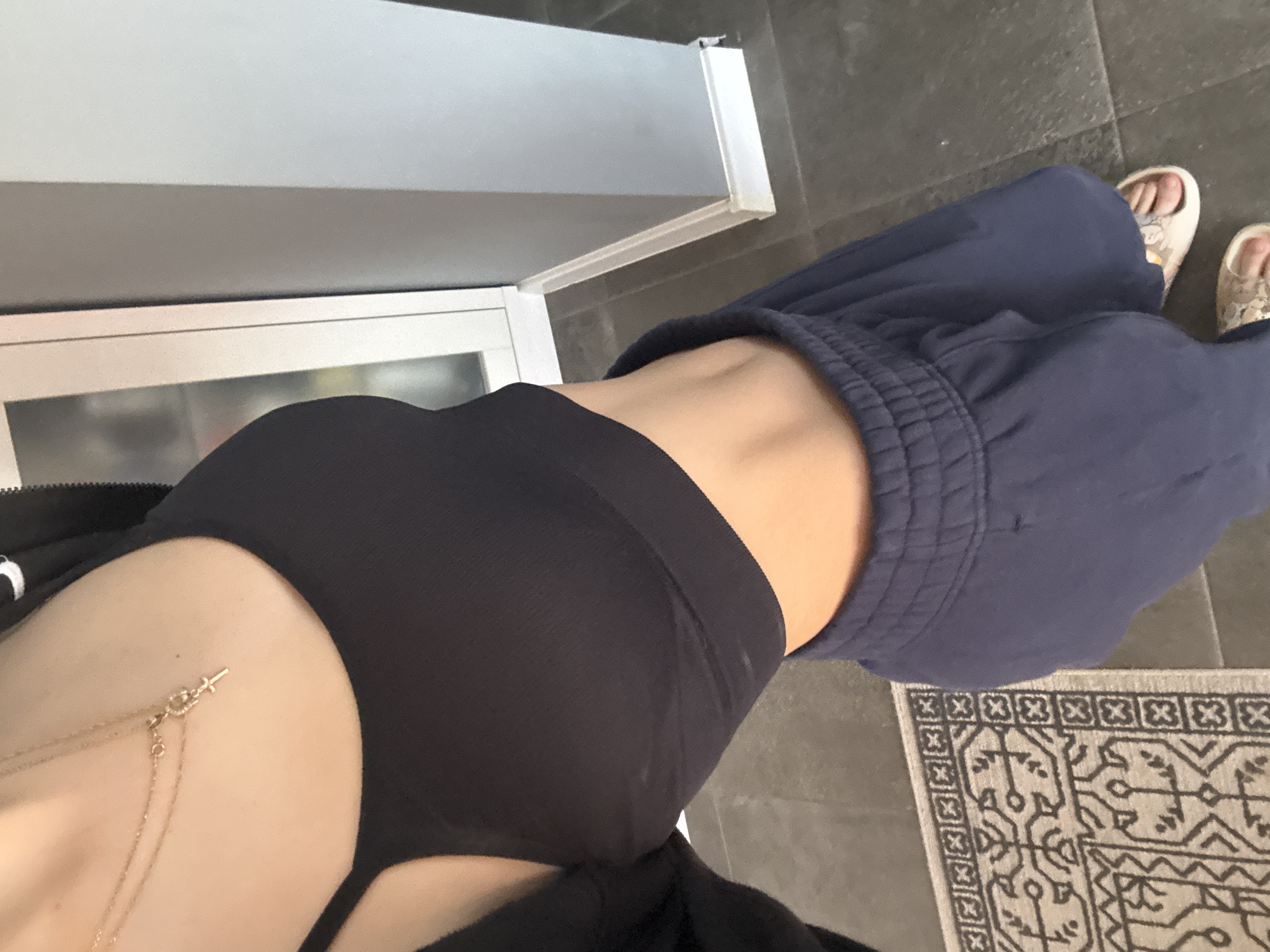 A top-down shot shows a person wearing black underwear and loose-fitting blue sweatpants, with visible skin on their midriff. A delicate gold necklace with a cross pendant adorns the person's upper chest. The individual is also wearing sandals. Part of a grey rug with a geometric pattern is visible on the right side of the image. The location cannot be determined.