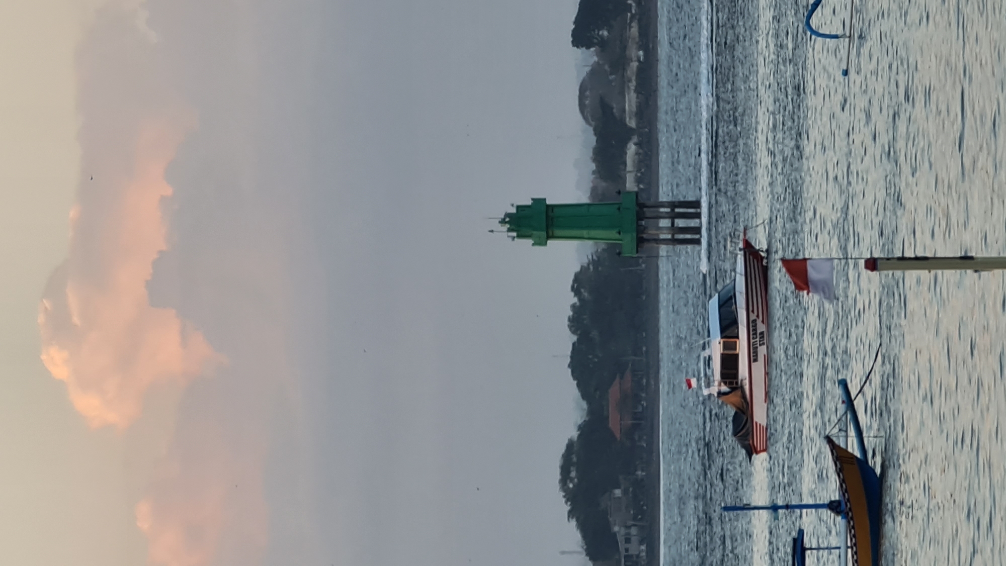 This media file depicts a serene maritime scene, likely at sunset, in Denpasar, Indonesia. The prominent subject is a green lighthouse, standing tall on pilings in the water. In the foreground, two boats are visible. The boat closer to the lighthouse is a sleek, modern vessel with white and red stripes, and bears the text "MARUTI CARGO STAR" on its side. A red and white flag, possibly the Indonesian flag, flies from a pole on this boat. Another, more traditional boat with a yellow and blue hull is partially visible in the foreground, its wooden structure suggesting local craftsmanship.
The water is calm, with gentle ripples reflecting the overcast sky. The sky itself is dramatic, with large, fluffy clouds tinged with pink and orange hues, indicating the soft light of either sunrise or sunset. In the distance, a treeline borders the water, with glimpses of buildings, possibly houses or resorts, nestled amongst the foliage. The overall mood is peaceful and tranquil, with the soft light and calm water contributing to a sense of quietude. The presence of the lighthouse and boats suggests a working harbor or a popular coastal area, while the natural beauty of the sky and water provides a picturesque backdrop.