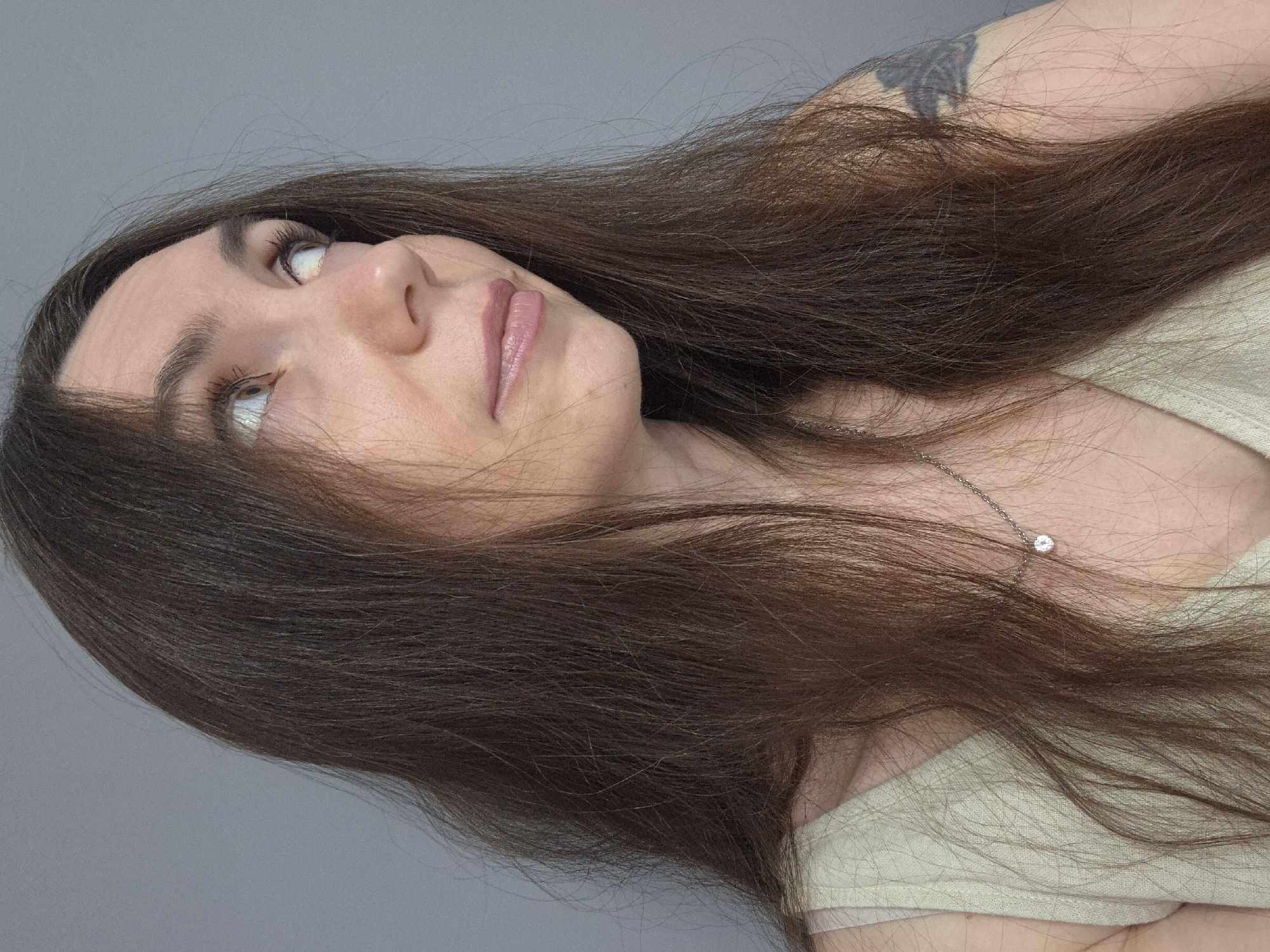 A medium shot shows a woman with long, dark brown hair that is parted on the left and falls over her right shoulder. She is looking to her right, with her head tilted slightly upwards. Her eyes are a light color, and her lips are painted a soft pink. She is wearing a pale, sleeveless top and a delicate silver necklace with a small pendant. A tattoo is visible on her upper left arm. The background is a solid, gray wall. The lighting is soft and even, casting no harsh shadows. The image appears to be a portrait taken indoors.