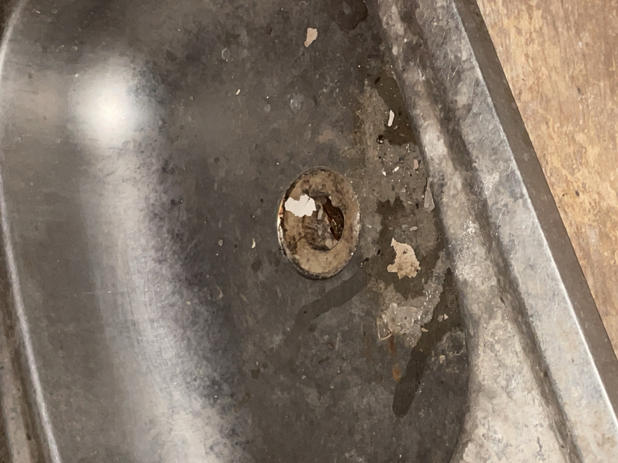 The image shows a close-up, overhead view of a dirty, stainless steel sink. The sink is stained and appears to have debris around the drain. The drain itself is partially covered with what looks like rust and peeling material. Water marks and some dried liquid are visible on the surface of the sink. The surrounding area, where the sink meets a countertop, is also visible and appears to be made of a light-colored wood-grained material. There are no people or explicit actions depicted in the image. The scene suggests a kitchen or bathroom sink in a state of disrepair or in need of cleaning. The location in Lokoja, Nigeria, is not visually represented in the image itself, but is provided as contextual information.