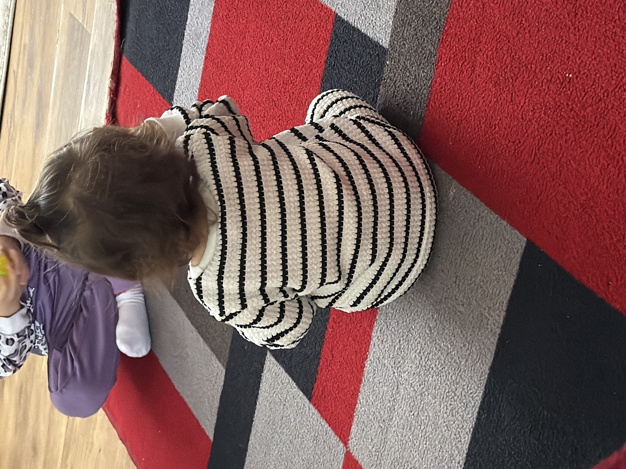 A child wearing a white and black striped sweater is sitting on a rug with red, grey, and black geometric patterns. The child has light brown hair. A second child wearing a white and black patterned long-sleeved shirt and purple pants is partially visible to the left, appearing to hold a yellow object. The scene is inside, likely on a wooden floor. Location information is not discernible from the image.