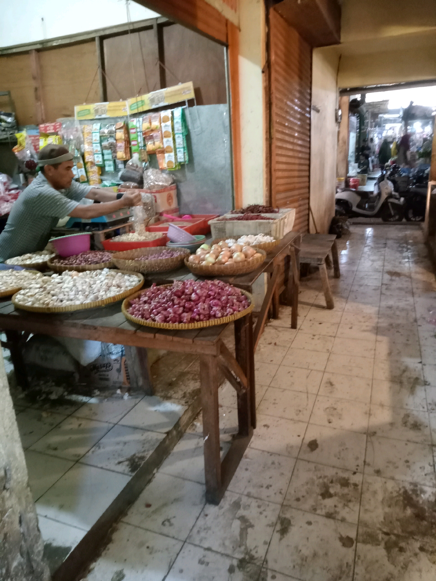 The image depicts a bustling market scene in Cirebon, Indonesia. A vendor, wearing a green and white striped shirt and a cap, is actively weighing goods on a scale, with his hands reaching towards a pile of what appears to be dried goods. In front of him, a wooden table is laden with various produce, including large trays of red onions, white onions, and other unidentifiable items, possibly shallots or garlic, arranged in woven baskets. Behind the vendor, shelves are stocked with packaged goods, likely snacks or food items, and a pink basin is visible. The market floor is tiled and appears to be somewhat dirty, with scattered debris. In the background, an open doorway reveals more market stalls and a glimpse of the street with parked motorcycles and people milling about, suggesting a lively commercial atmosphere. The architecture features wooden structures and a large, rolled-down shutter, characteristic of traditional Indonesian markets.