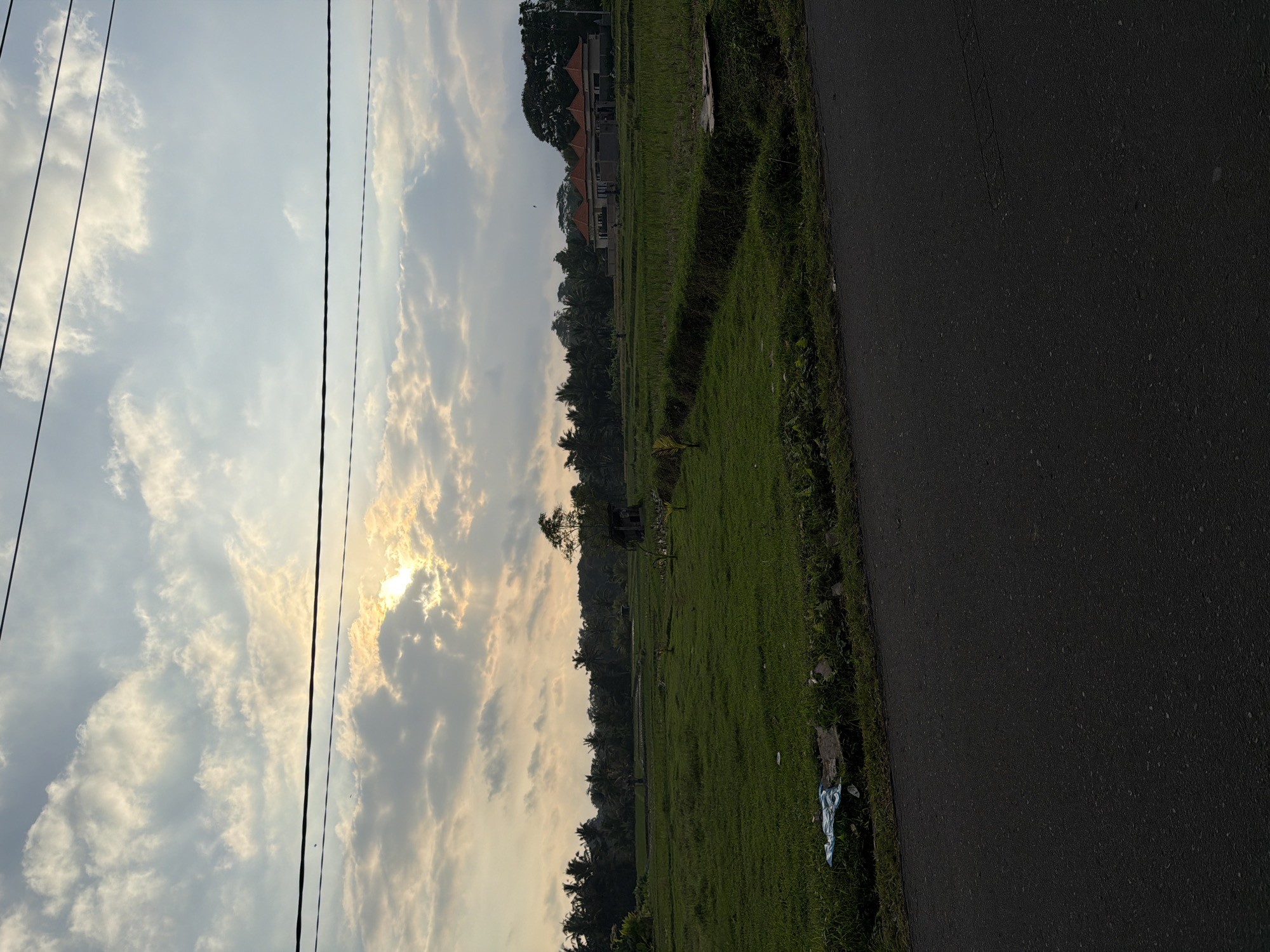 The image captures a rural landscape at dusk. The sky is filled with a mixture of clouds, with the sun peeking through them, casting rays of light. In the foreground, a dark asphalt road runs horizontally across the bottom of the frame. To the left of the road, there is a patch of green grass with some debris, including a blue plastic bag. The middle ground features a lush green rice field, with a ditch running through it. In the distance, a line of palm trees and other vegetation borders the field. A modern house with orange tiled roofs is visible on the right side of the field, surrounded by trees. The overall impression is one of a serene, rural setting transitioning from day to night.