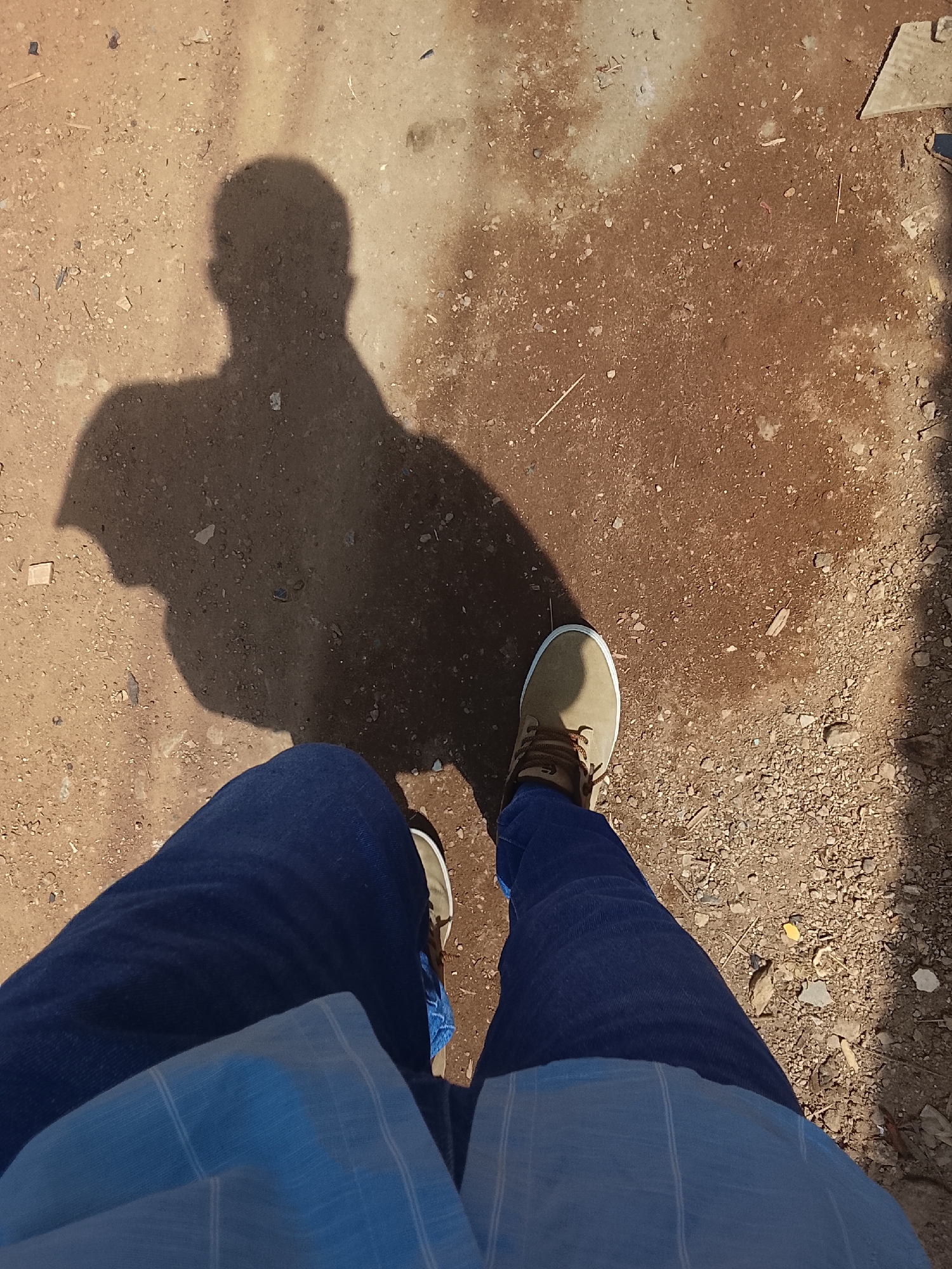 The photograph shows a person's perspective looking down at their feet and shadow on a dirt and gravel ground. The person is wearing blue jeans and tan shoes with brown laces. A light blue striped shirt is visible. The shadow suggests the person is facing the sun. The ground is uneven with varying shades of brown dirt and scattered gravel. Based on the environment it is difficult to pinpoint a specific location within Abuja, Nigeria, but it appears to be an outdoor area with natural ground cover. There is no clear evidence of other people or significant objects. The main action is the person standing on the ground. No interaction is visible.