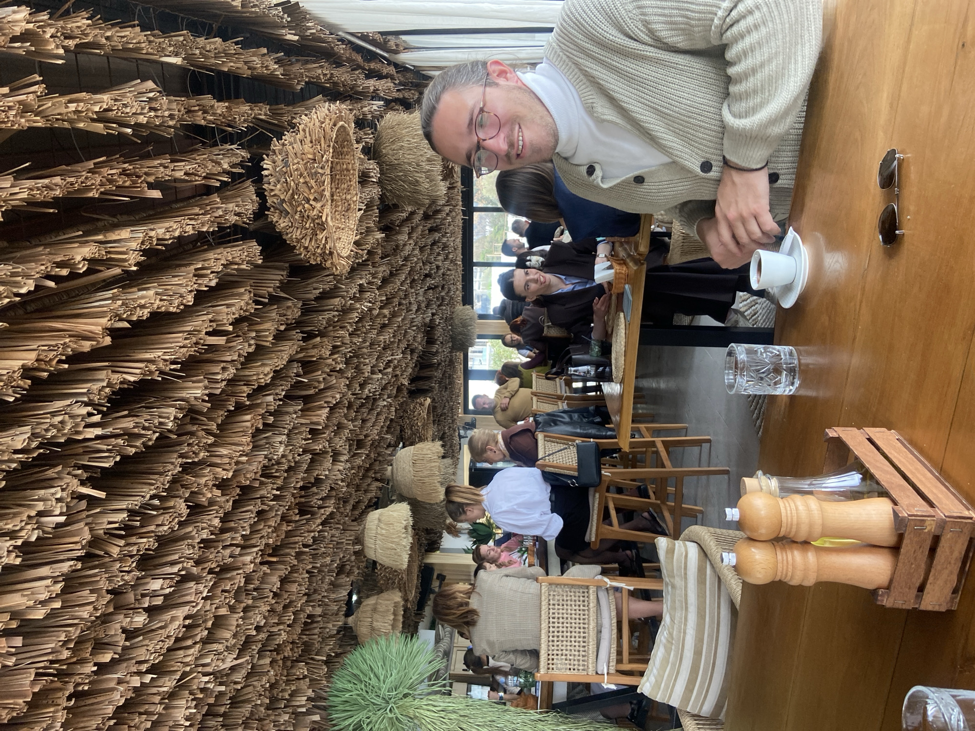 The image depicts the interior of a restaurant or cafe located in Petrovac centar, Montenegro. The ceiling is entirely covered with numerous hanging, dried, light brown natural fibers or strips, creating a thatched aesthetic. Several large, woven basket-like light fixtures or decorative elements are suspended from this ceiling.
In the foreground, a light-skinned male, appearing to be in his 30s or 40s, with light brown hair pulled back, is seated at a light-colored wooden table. He wears clear-rimmed eyeglasses, a white turtleneck shirt, and a light beige knitted cardigan with dark buttons. He is smiling and looking towards the viewer. His right hand rests on a small white cup and saucer containing a dark liquid, likely coffee. On the table directly in front of him are the cup and saucer, a clear glass of water, and a pair of dark sunglasses with a thin silver bridge. To his left, a wooden caddy holds two wooden grinders (salt and pepper shakers) and a clear bottle, likely containing olive oil. A striped cushion (beige and white) is visible on a chair in the immediate foreground.
In the mid-ground and background, numerous other patrons are seated at similar wooden tables and chairs or high stools. To the left, a woman with brown hair (viewed from behind) wears a light brown patterned jacket and sits on a woven chair. Another woman, further back and to the main subject's left, wears a dark jacket and smiles. A man in a light brown sweater is visible standing or seated in the right background. The establishment appears well-lit by natural light entering through large windows or open sections along the back wall, which offer a glimpse of an indistinct outdoor area, possibly a promenade or waterfront. Green foliage, resembling large palm fronds or artificial plants, is visible on the left side of the frame.