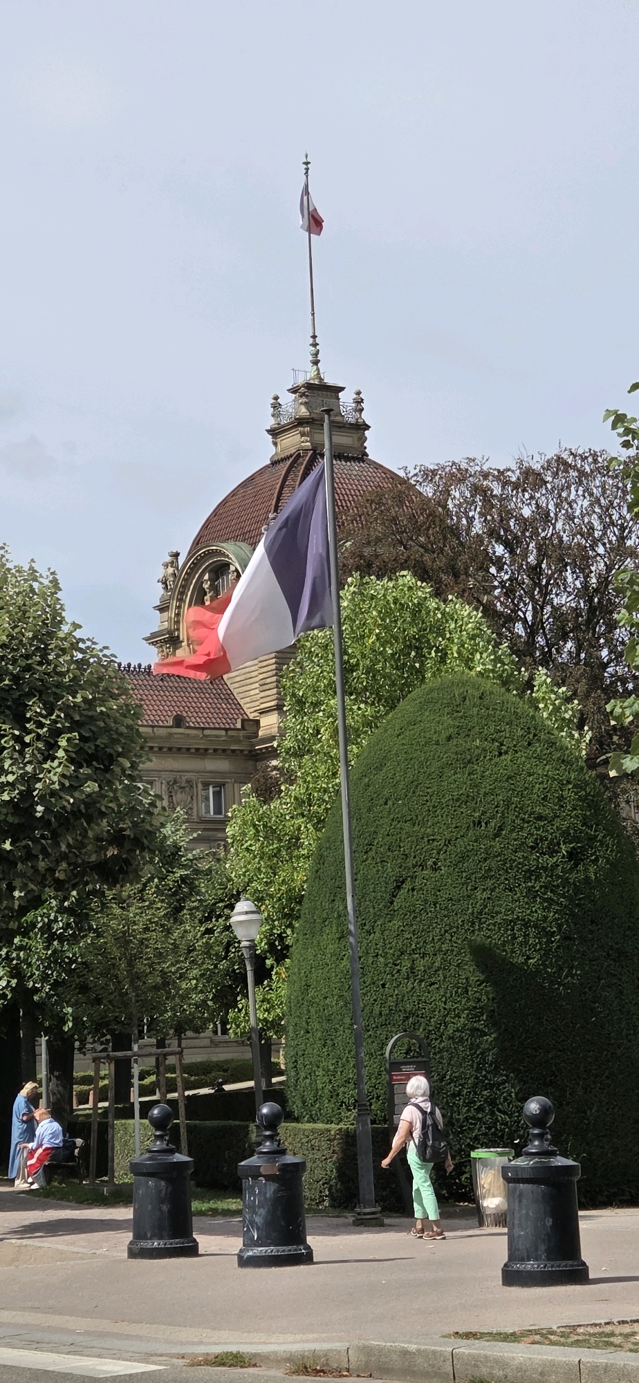 The image depicts a view in Strasbourg, France, with the Strasbourg Cathedral's dome and a portion of its facade visible in the background. A French flag flies prominently from a flagpole in the foreground. The scene includes lush green trees and manicured bushes framing the view. In the middle ground, several black bollards are placed along a paved walkway. To the left, two individuals are seated on a bench, one wearing a blue garment and the other a red and white striped shirt. To the right, a woman with white hair wearing a light pink shirt and light green pants walks away from the camera, carrying a backpack. A lamppost is visible behind the bollards. The sky is overcast.