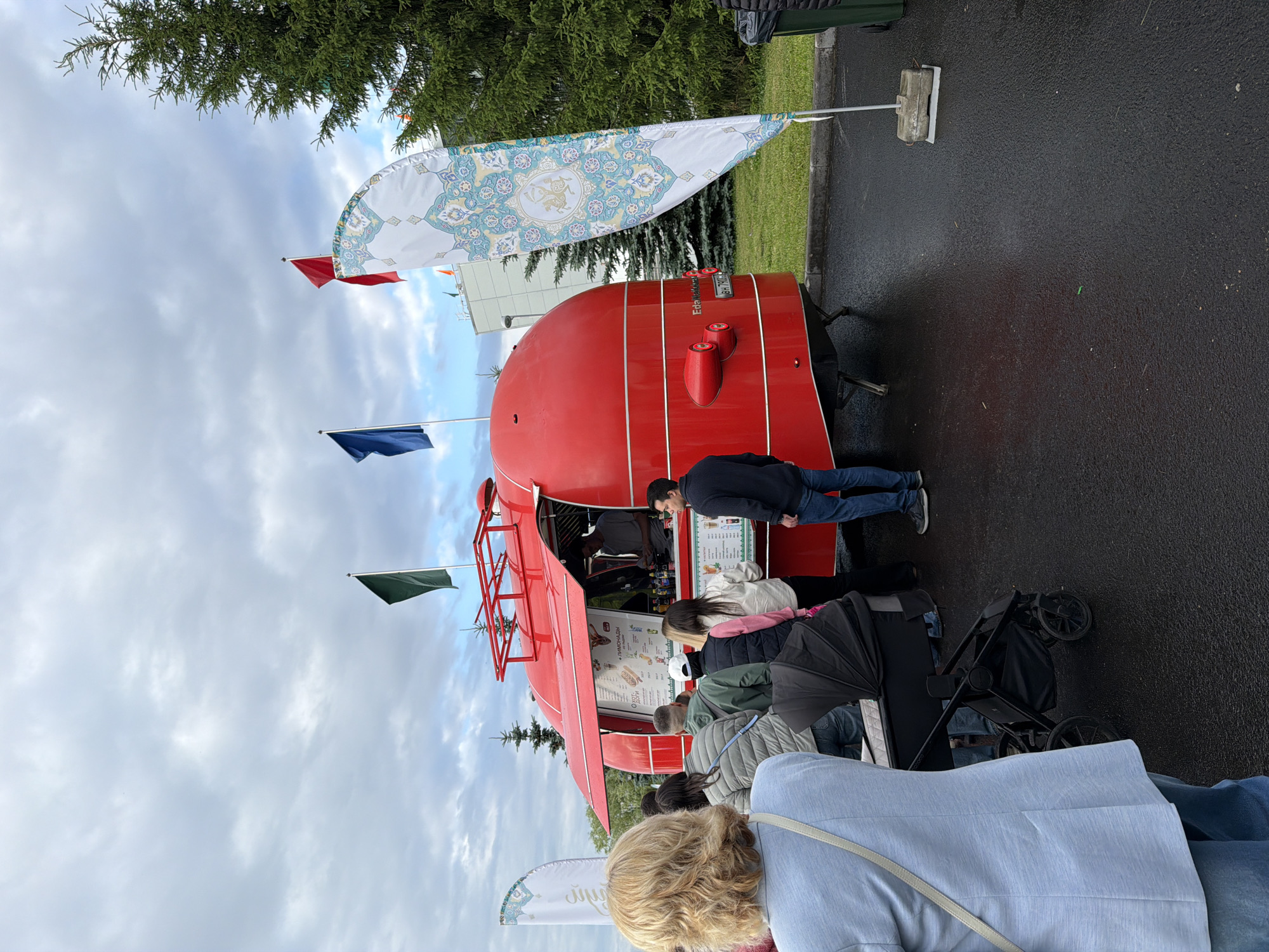 An outdoor scene features a bright red, rounded food truck with a red awning open for business. Several people are standing in line, facing the truck, while one person appears to be serving customers from inside. The ground is wet, suggesting recent rain. Flags of various colors, including blue, green, and red, are visible, along with a decorative flag featuring a circular, mandala-like pattern. A large evergreen tree is situated behind the food truck. A stroller is parked near the line of people. The sky is overcast with clouds. The scene appears to be a public gathering or event.